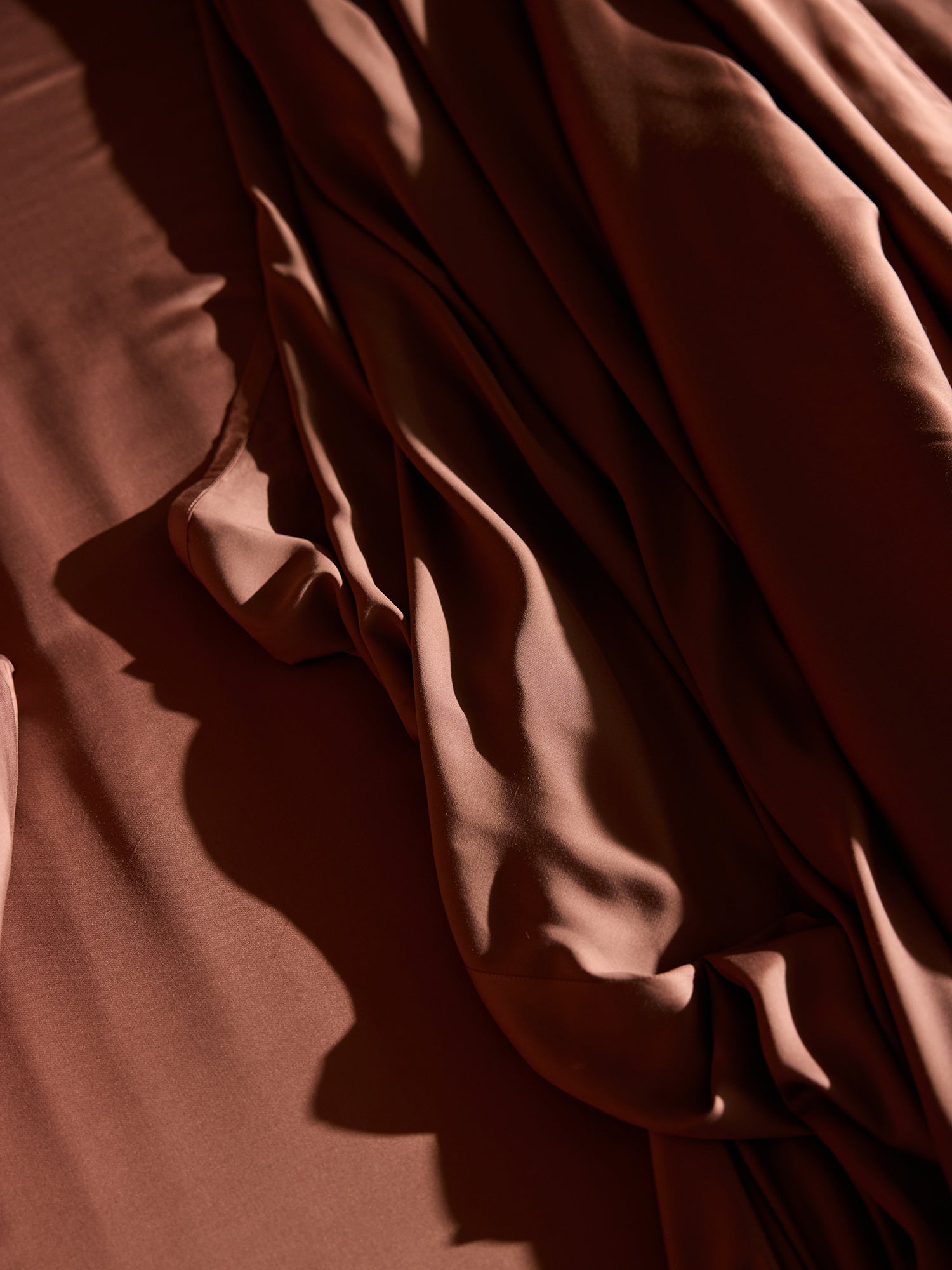 The Bamboo Flat Sheet - Wholesale by Cozy Earth exhibits a rich, brown satin appearance, artfully draped and softly illuminated by gentle light, creating smooth folds and shadows. The silky, luxurious texture of the material reflects light to emphasize its sheen and fluidity. 