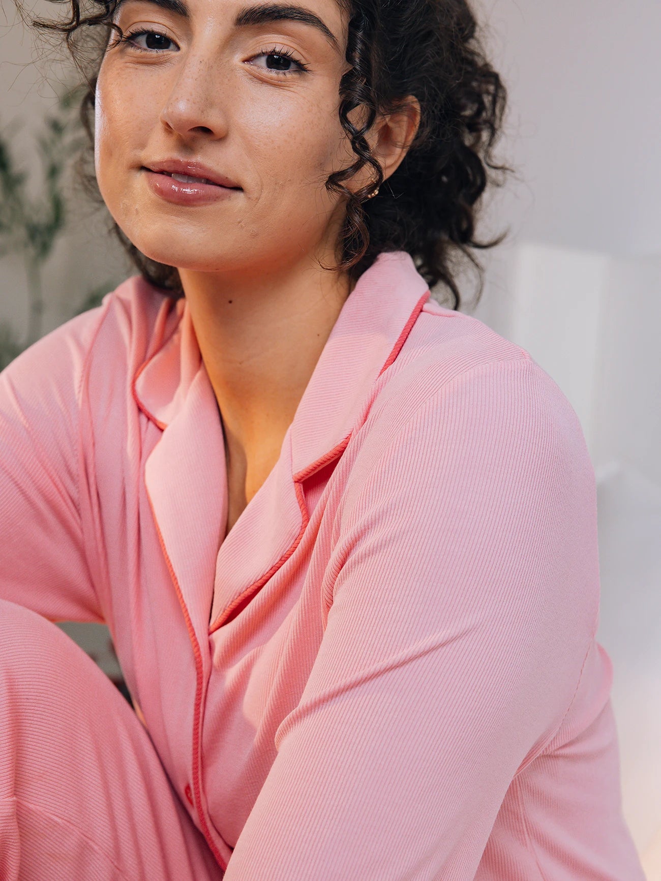 A woman with curly hair, wearing a Cozy Earth Women's Bamboo Rib Knit Classic Long Sleeve Pajama Top, sits comfortably indoors. The soft lighting creates a warm atmosphere. They wear a gentle expression, and green plants blur in the background. 