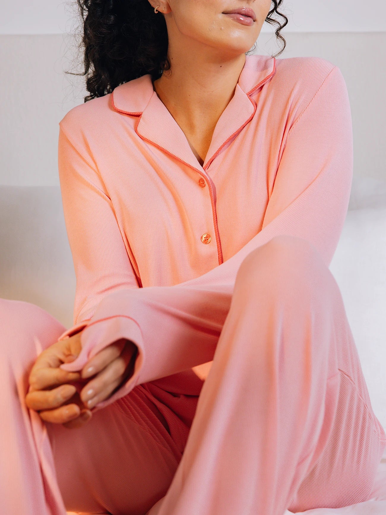 A woman basking in soft light wears Cozy Earth's Women's Bamboo Rib-Knit Classic Pajama Pant in pink, with a collar and buttons. They have gently clasped hands and curly hair, with a white pillow and wall behind them. 