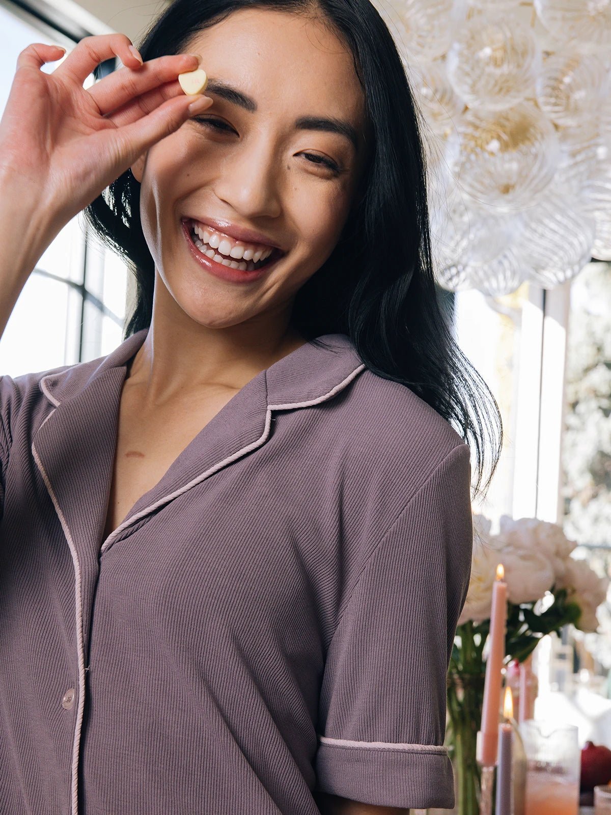 A smiling woman with long dark hair holds a small item near their eye, wearing Cozy Earth's Women's Bamboo Rib-Knit Classic Short Sleeve Pajama Top. In the background, lit candles and white flowers adorn a table. 