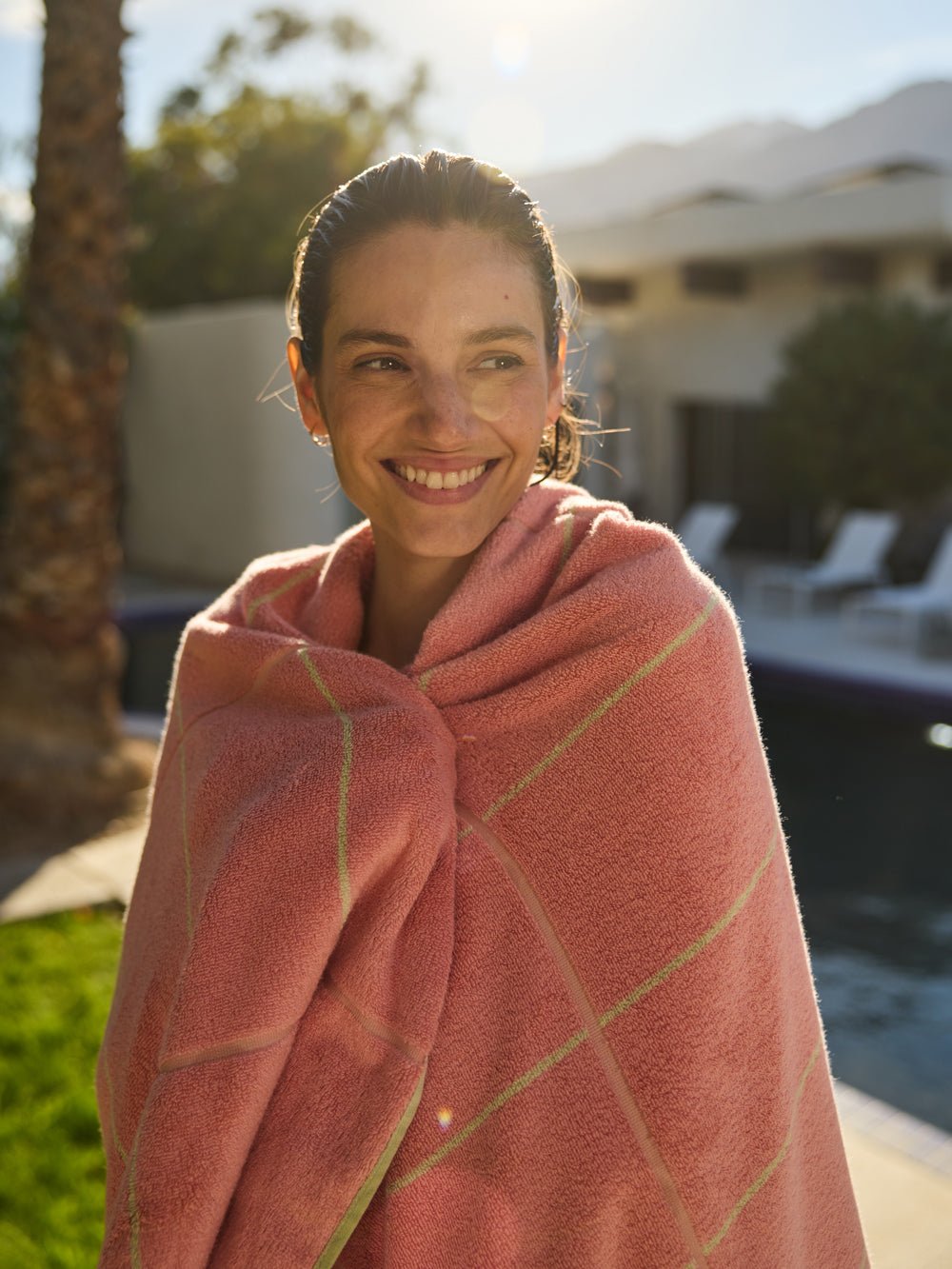 A person smiles while wrapped in a Cozy Earth Windowpane Resort Towel, its pink checkered pattern catching the sunlight. They stand outside by a pool with their face lit up, surrounded by palm trees, grass, and a building in the background. 