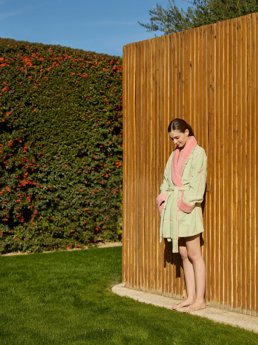 A woman wearing the Cozy Earth Windowpane Resort Robe in green with pink trim stands barefoot on grass, leaning against a wooden fence. A tall hedge with red flowers decorates the background beneath a clear blue sky. 