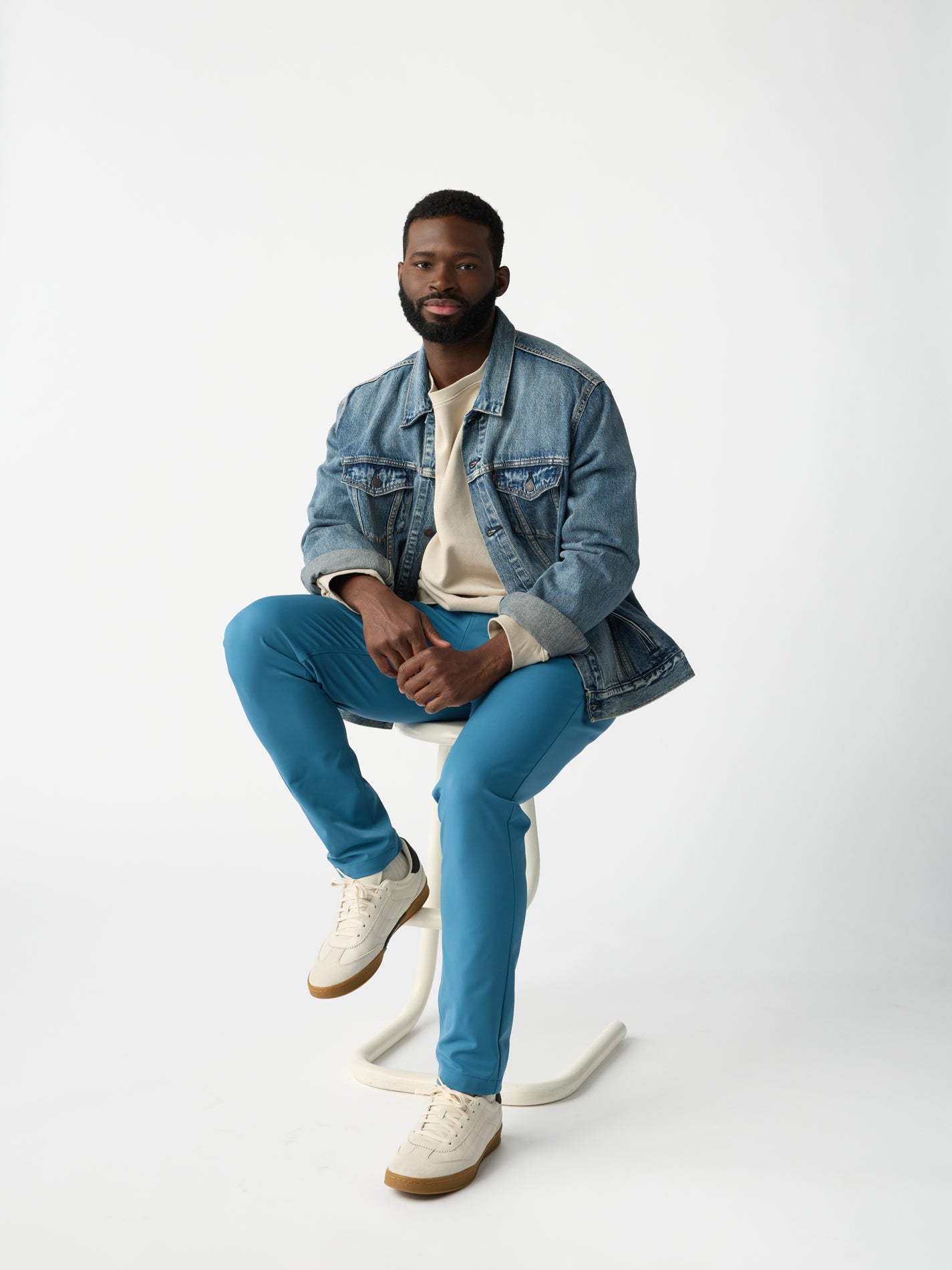 A man is seated on a modern stool against a white background, wearing a denim jacket, light shirt, and Cozy Earth Men's Everywhere Pant in bright blue, paired with white sneakers. The look is relaxed and casual. 