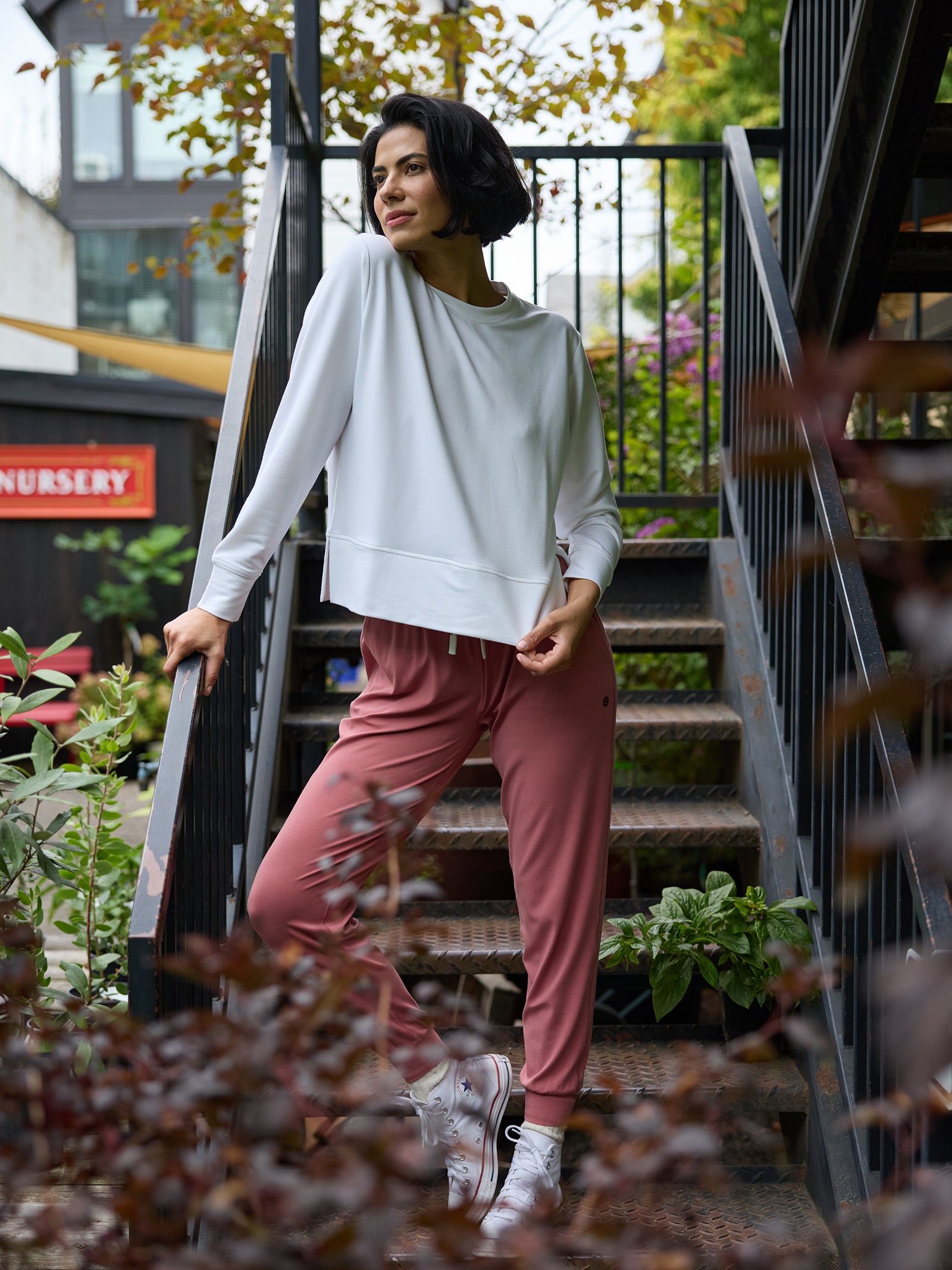 A person with short dark hair stands on an outdoor staircase, wearing a white Women's StudioLite Crewneck top from Cozy Earth and pink jogger pants. They have one hand resting on the railing and appear to be looking off into the distance. The surrounding area is lush with greenery. 