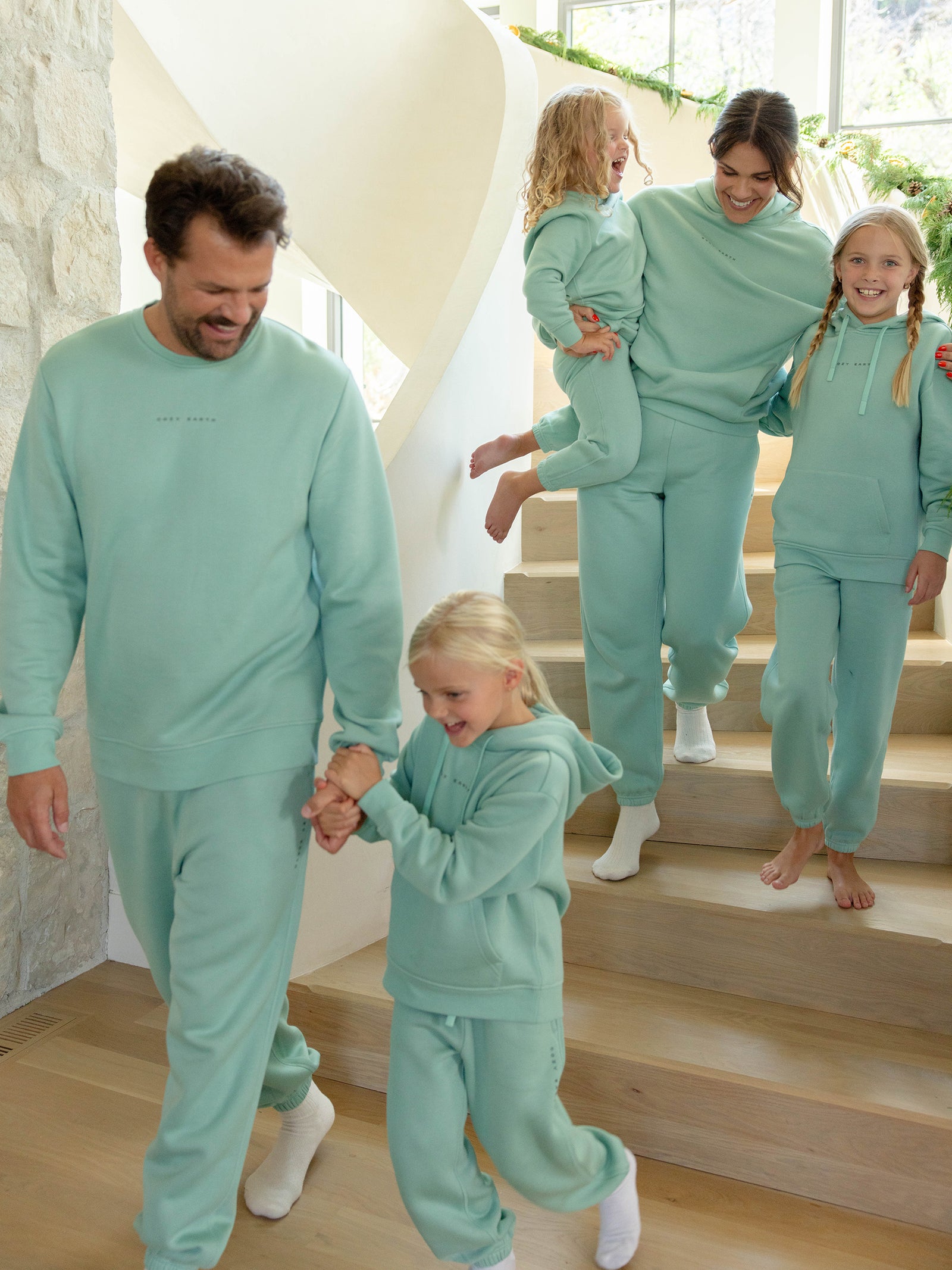 A family of five, all wearing matching Men's CityScape Crewnecks from Cozy Earth, cheerfully descends a wooden staircase. One adult carries a young child, while the others walk hand in hand. The space is filled with bright, natural light. 