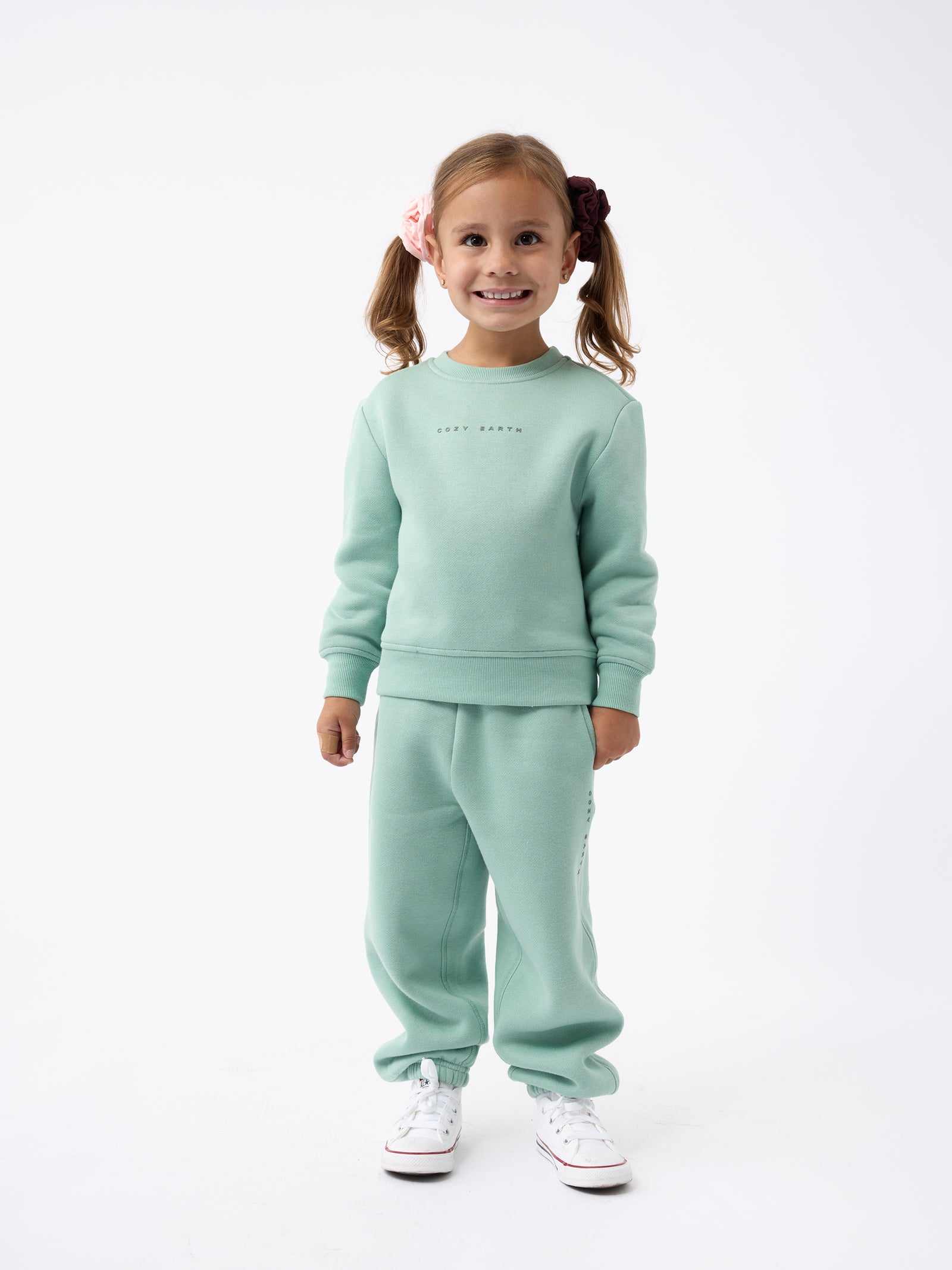 A young girl with pigtails stands smiling, dressed in the Kid's CityScape Crewneck by Cozy Earth and matching pants, paired with white sneakers against a plain white background. 