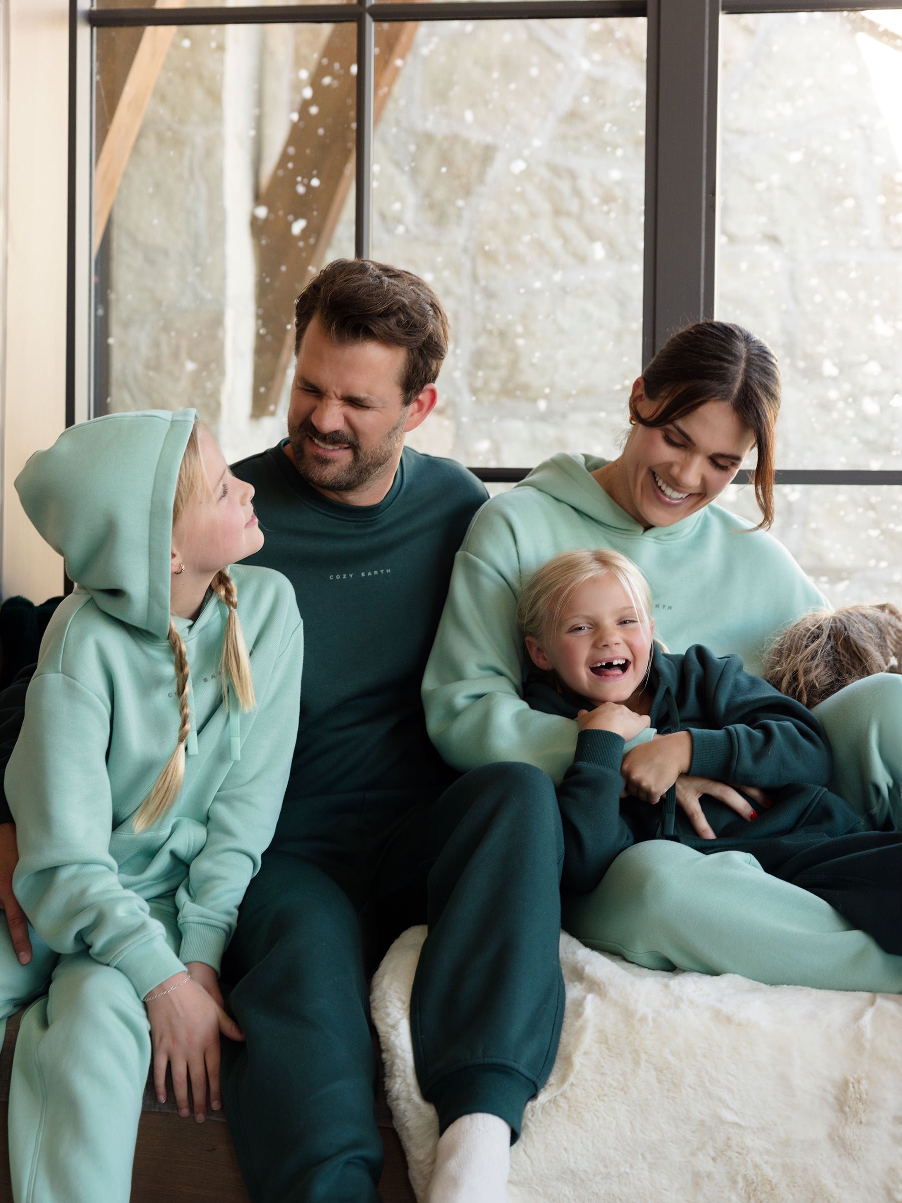 A family of four is gathered indoors, smiling as they wear matching teal and green Women's CityScape Hoodies from Cozy Earth, paired with sweatpants. The cozy and cheerful scene is complete with a snowy landscape visible through the large windows in the background. 