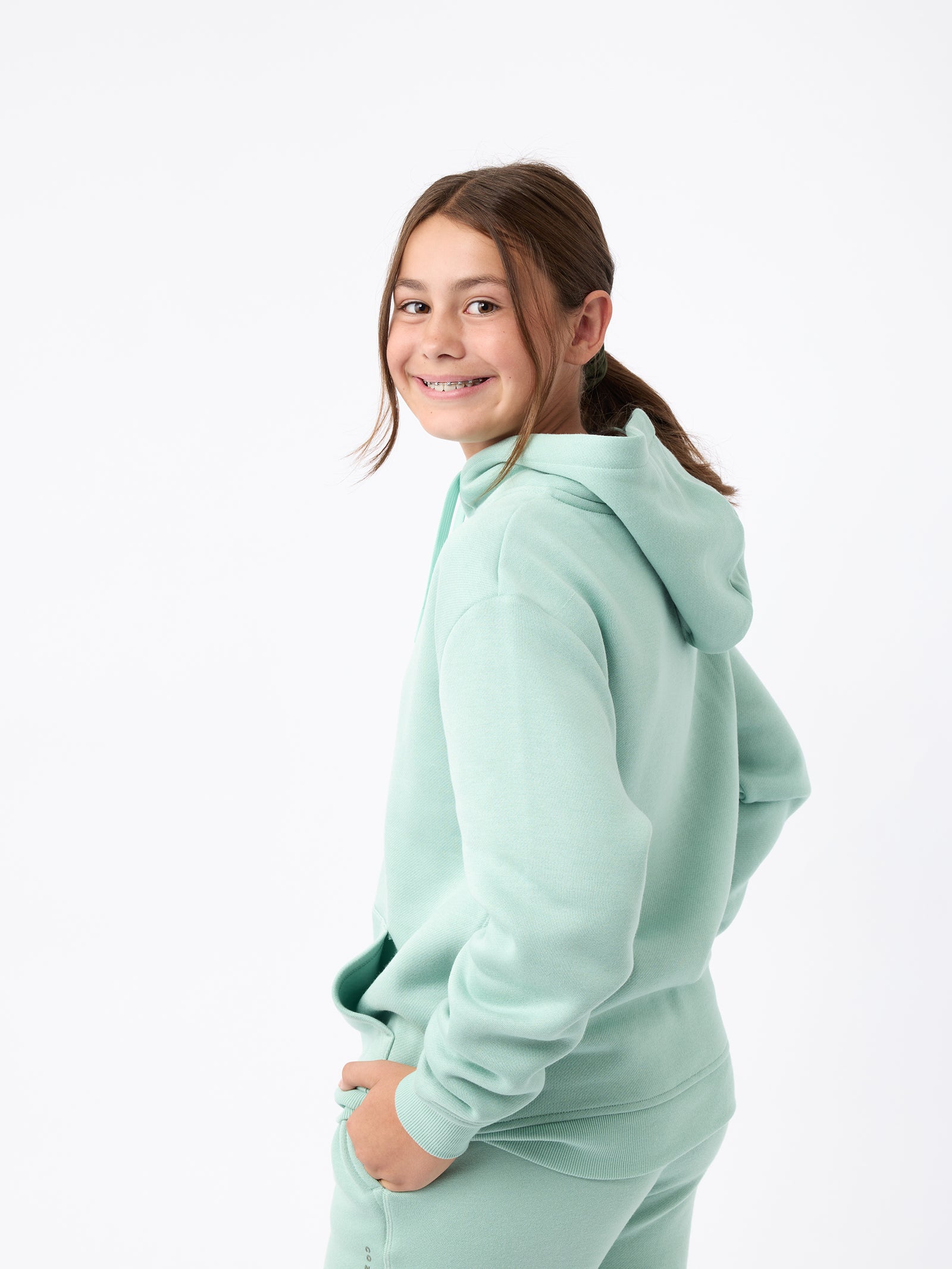 A young girl, dressed in a Cozy Earth Kid's CityScape Hoodie and matching pants in mint-green, smiles at the camera with her hair tied back in a ponytail. She stands against a plain white background. 