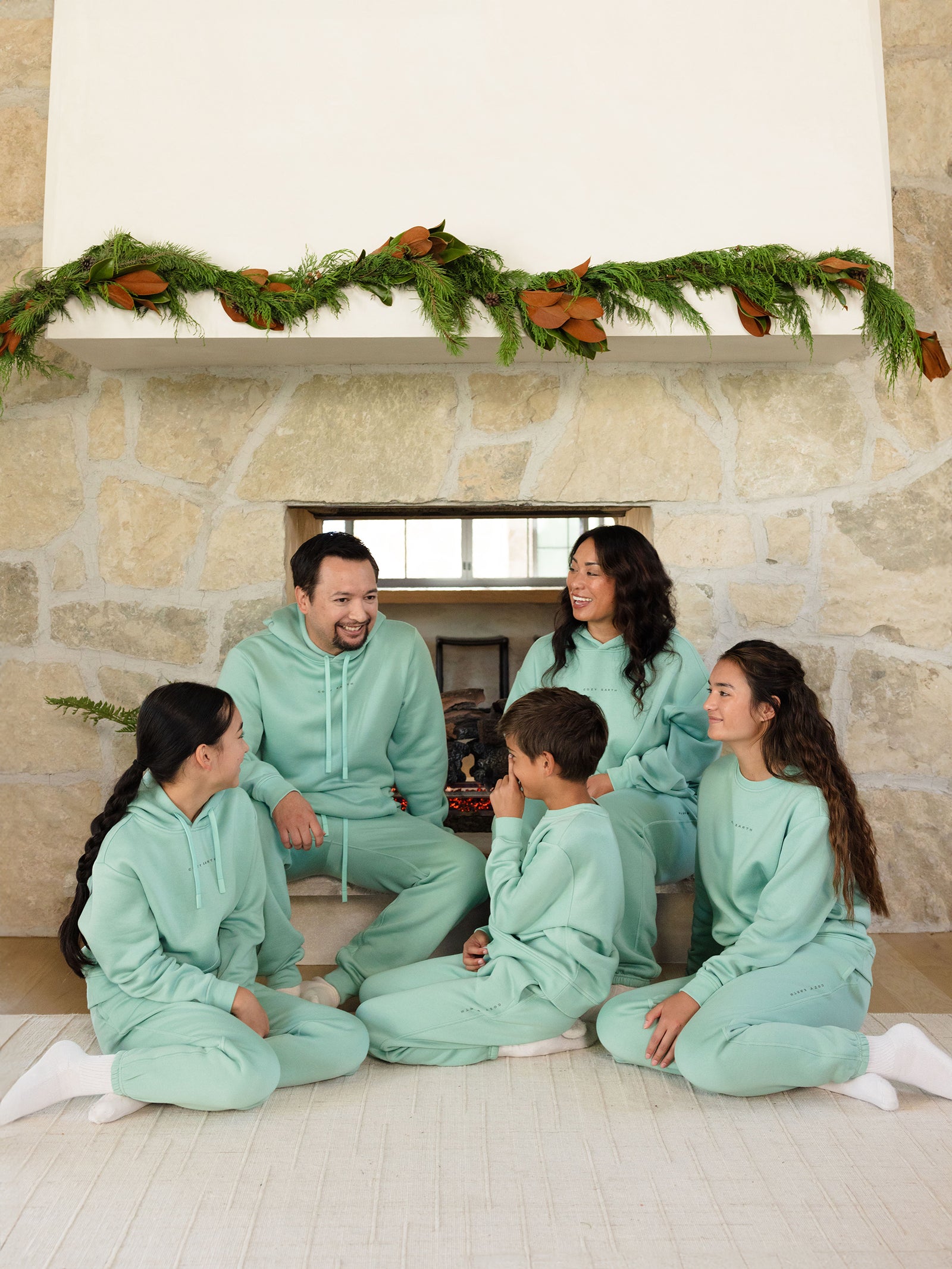 A family of five, dressed in matching mint green outfits including the Women's CityScape Sweatpant from Cozy Earth, sits in front of a fireplace adorned with a garland. They are sitting on a light rug, smiling and looking at each other. The room has stone walls and a warm ambiance. 