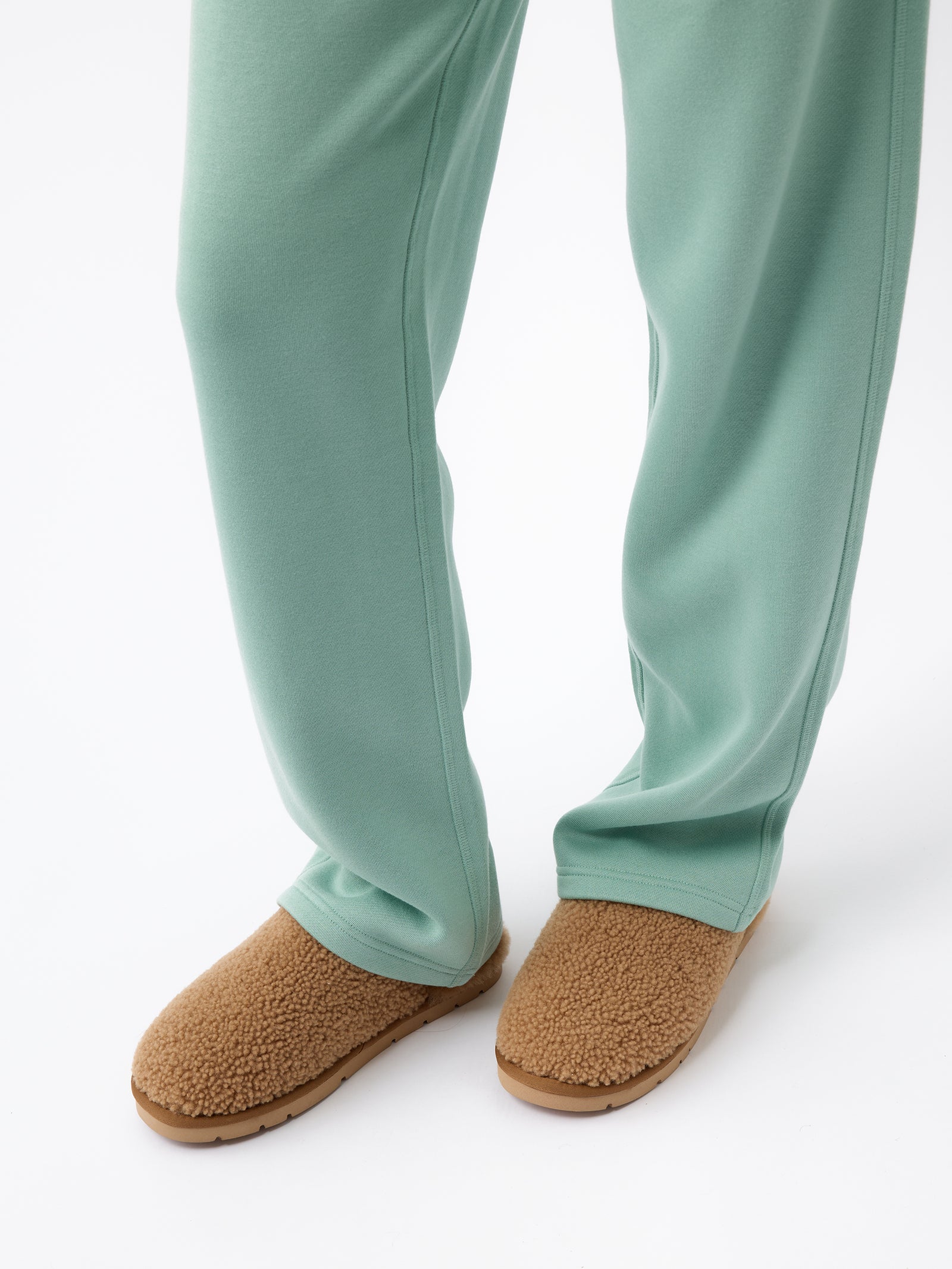 A person dressed in Cozy Earth's light green Women's CityScape Wide Leg Pant and brown sherpa-lined slippers is standing against a white background. The pants fully cover the person's legs, while the slippers have a soft, fuzzy look. 