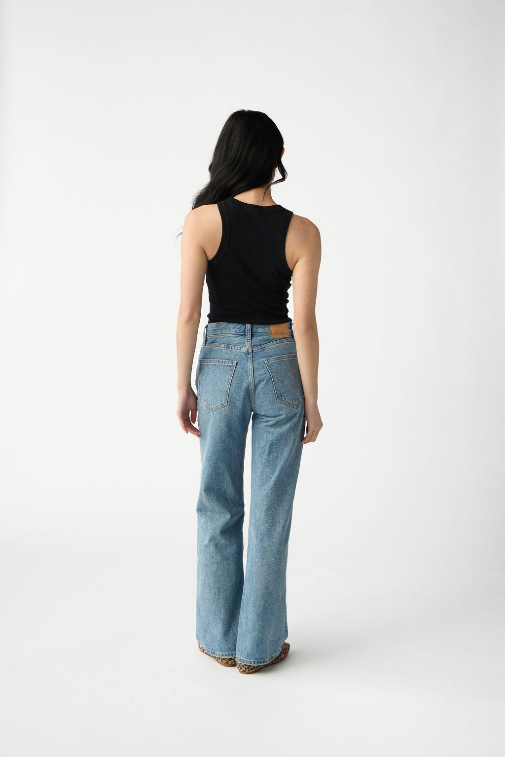 A dark haired woman wears Cozy Earth Women's Fine Ribbed Tank in Jet Black. She is facing away from the camera and stands in front of a white background. 