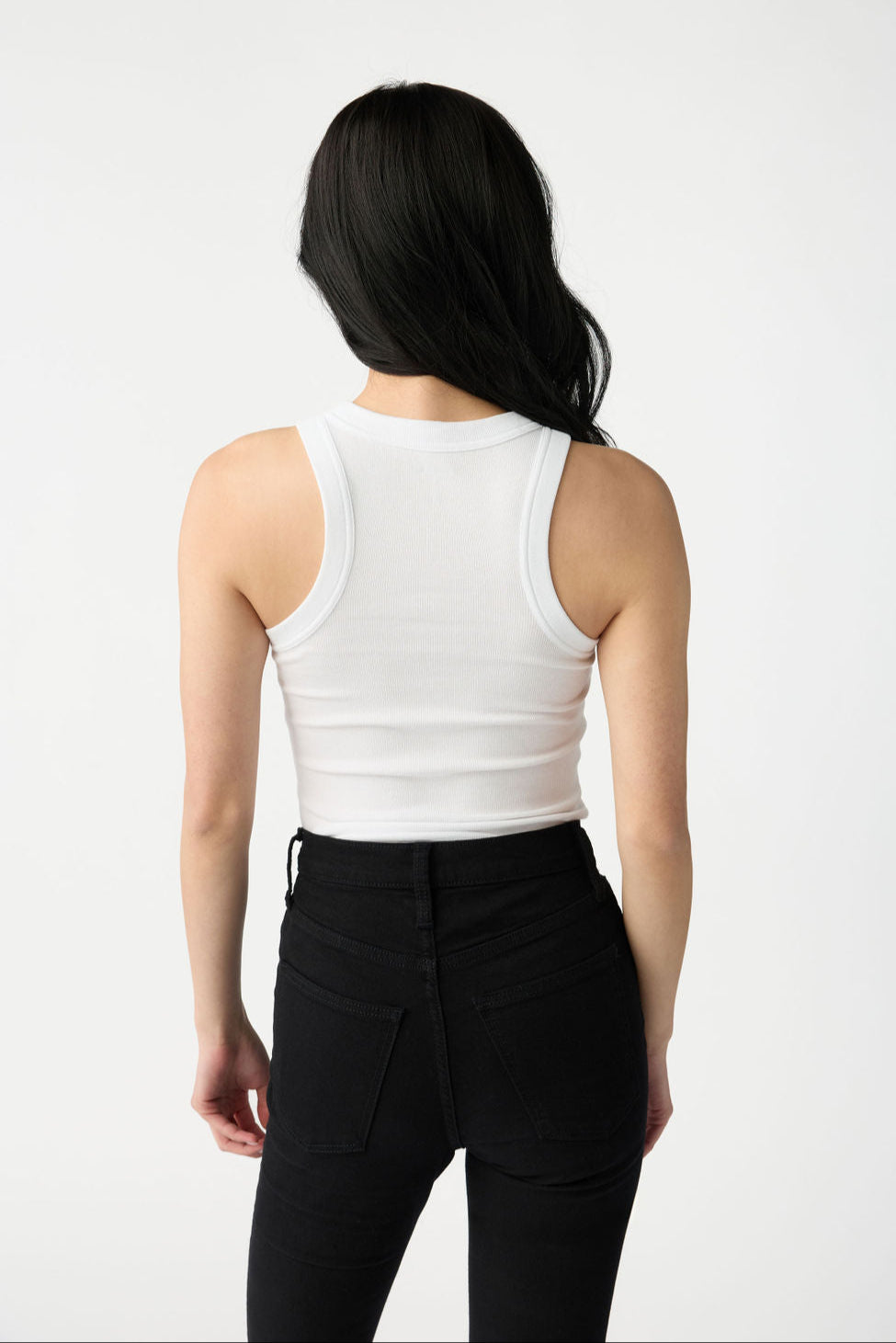 A dark haired woman wears Cozy Earth Women's Fine Ribbed Tank in White. She wears black jeans, faces away from the camera, and stands in front of a white background. 