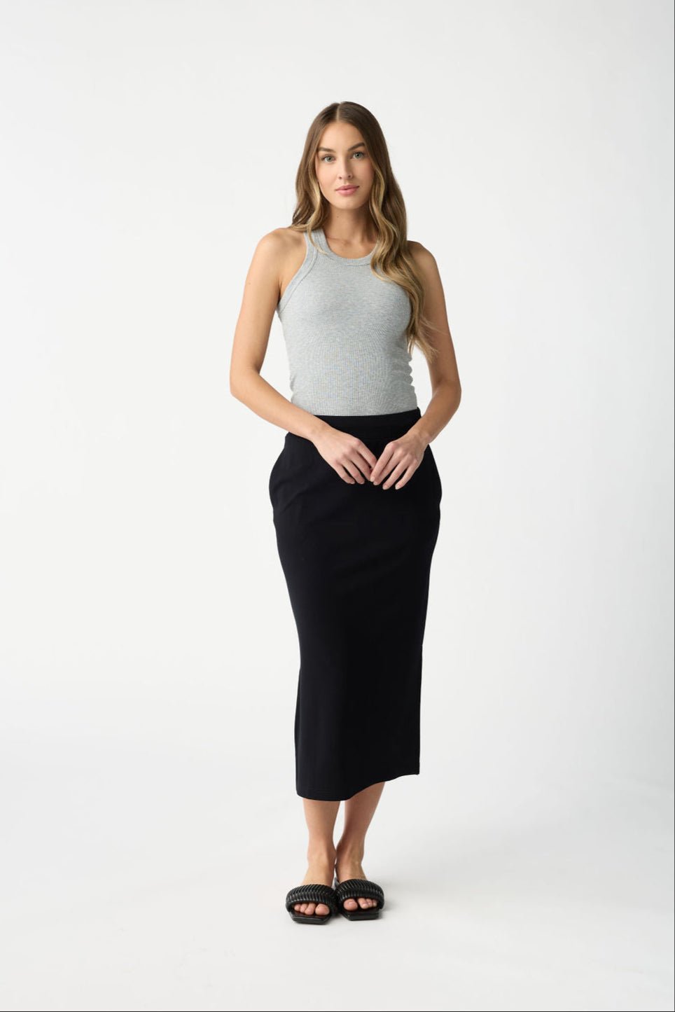 A woman with long wavy hair stands against a white background, wearing a gray sleeveless top, black sandals, and the Women's Brushed Bamboo Midi Skirt by Cozy Earth. Her hands rest in front of her. 