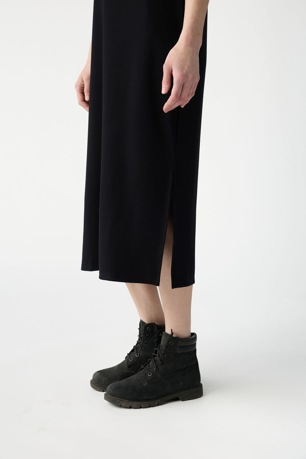 A person stands on a white background wearing a black midi dress and rugged boots, with the focus on the lower body and hands resting by their sides. Products: Women's Brushed Bamboo Sleeveless Midi Dress by Cozy Earth. 