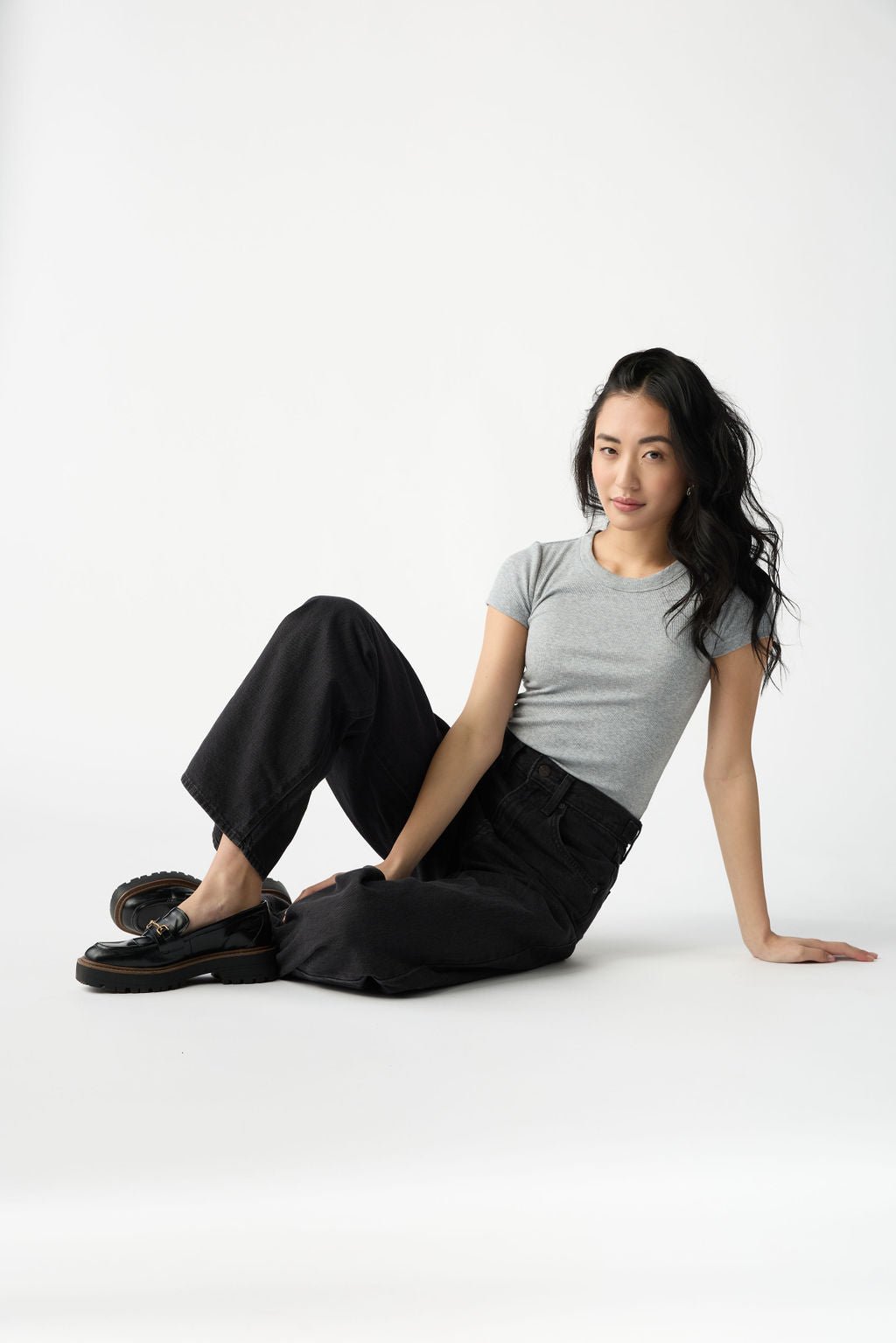 A dark haired woman wears Cozy Earth Women's Fine Ribbed Tee in Heather Grey. She is wearing black jeans and sits on the floor in front of a white background. 