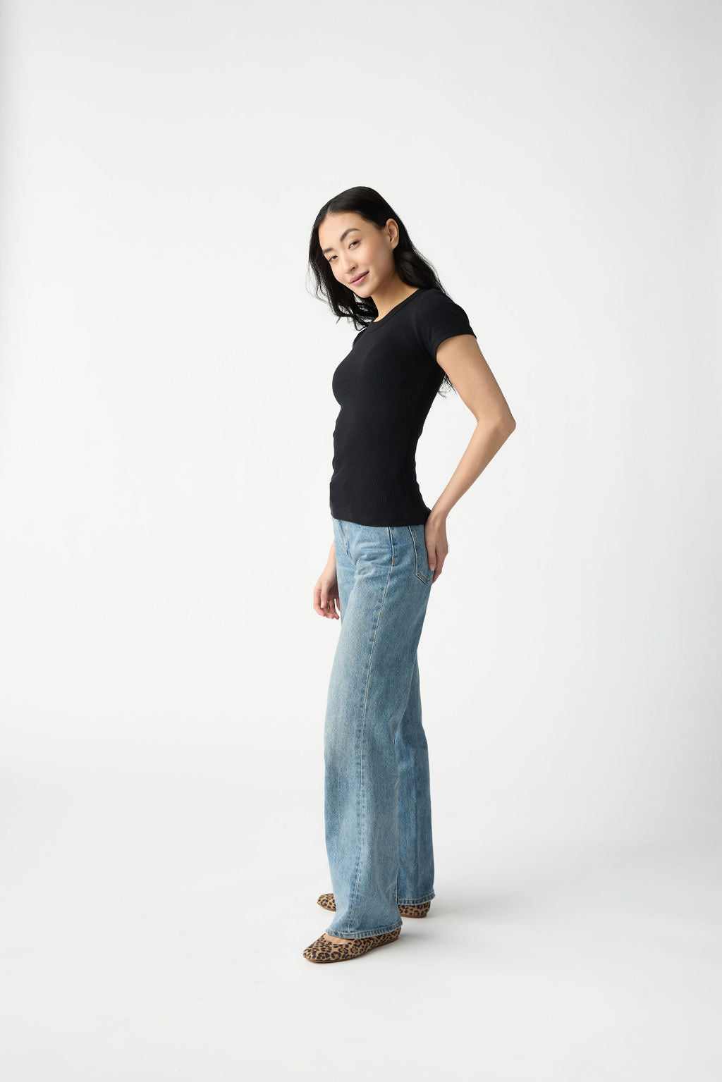 A dark haired woman wears Cozy Earth Women's Fine Ribbed Tee in Jet Black. She is wearing blue jeans and stands in front of a white background. 