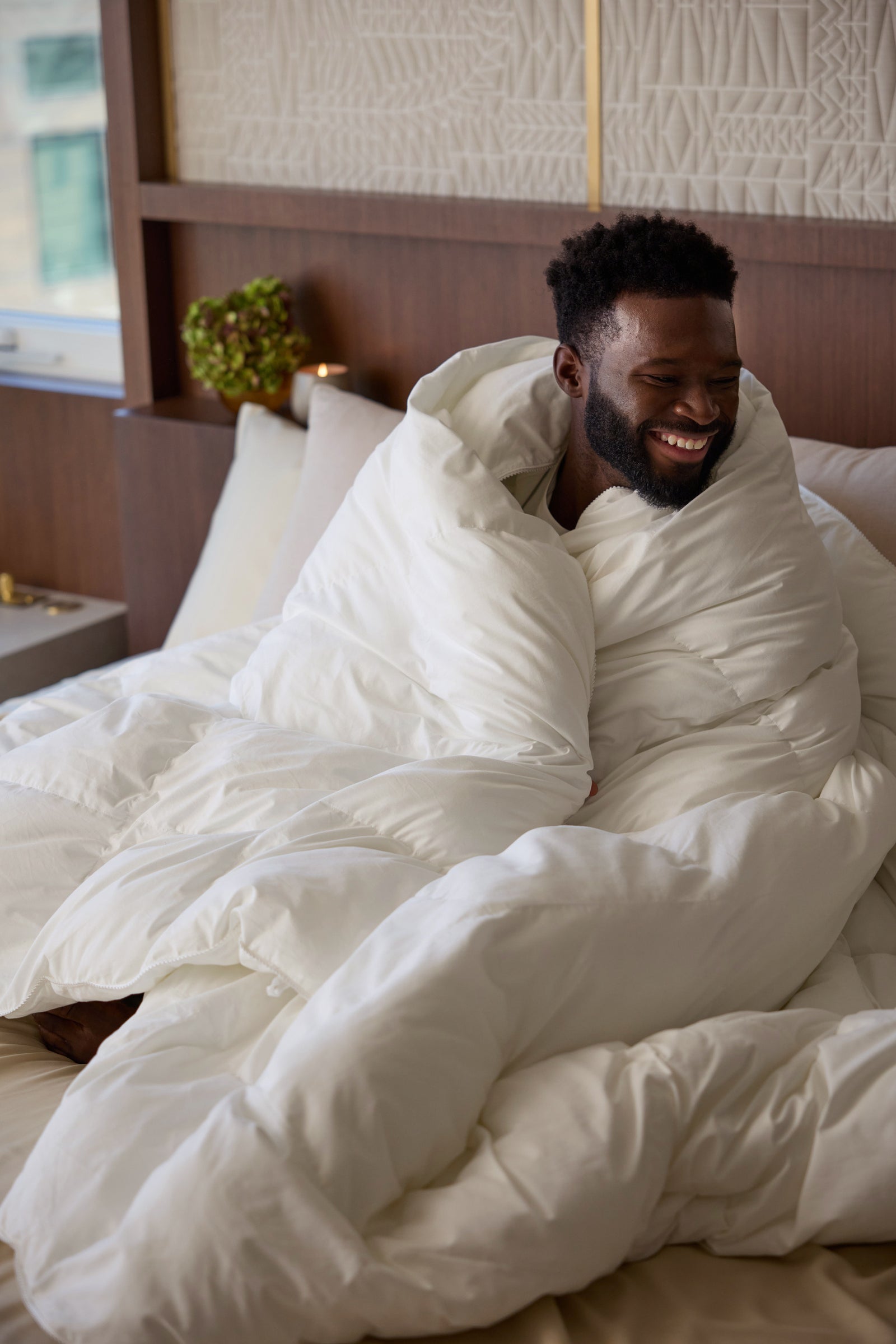 A man sits on a bed wrapped in the fluffy Cozy Earth Down Alternative Comforter, smiling and looking to the side in a cozy, well-lit bedroom. 