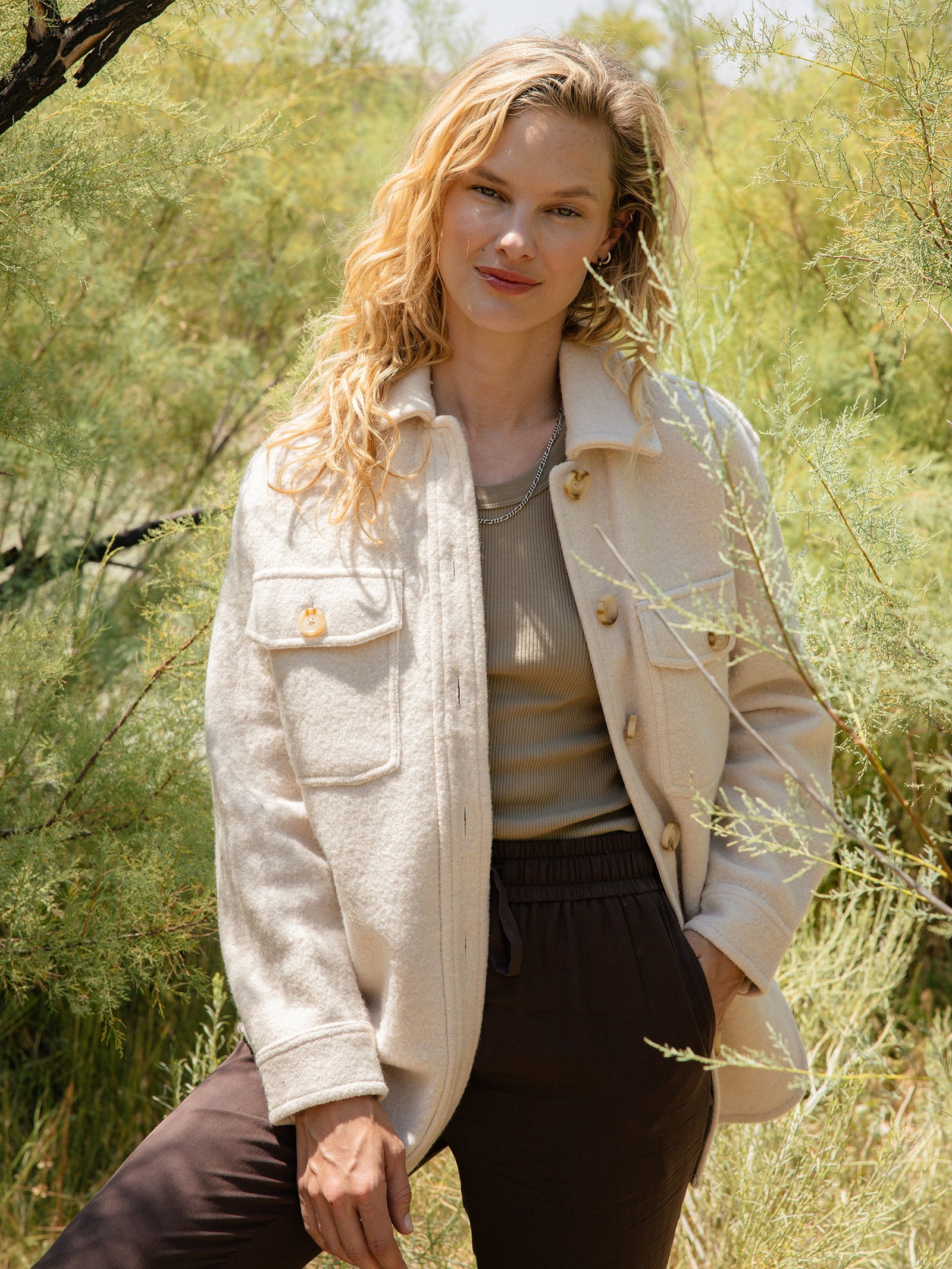 A blonde woman stands outdoors in front of greenery, wearing a light-colored Women's Boucle Shacket by Cozy Earth over a beige top and dark pants. Her wavy hair cascades over her shoulders, and she smiles gently while resting one hand against the side of her shacket.  