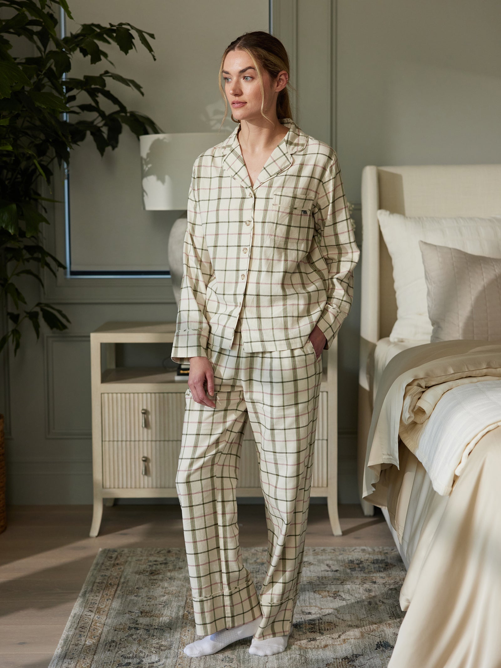 A person in the Women's Farmhouse Long Sleeve Pajama Set by Cozy Earth, featuring a soft white and green plaid pattern, stands beside a bed in a well-lit bedroom. The room exudes a calm atmosphere with light-colored bedding, a wooden nightstand equipped with a lamp, and some greenery in the background. The person has one hand resting in their pocket. 