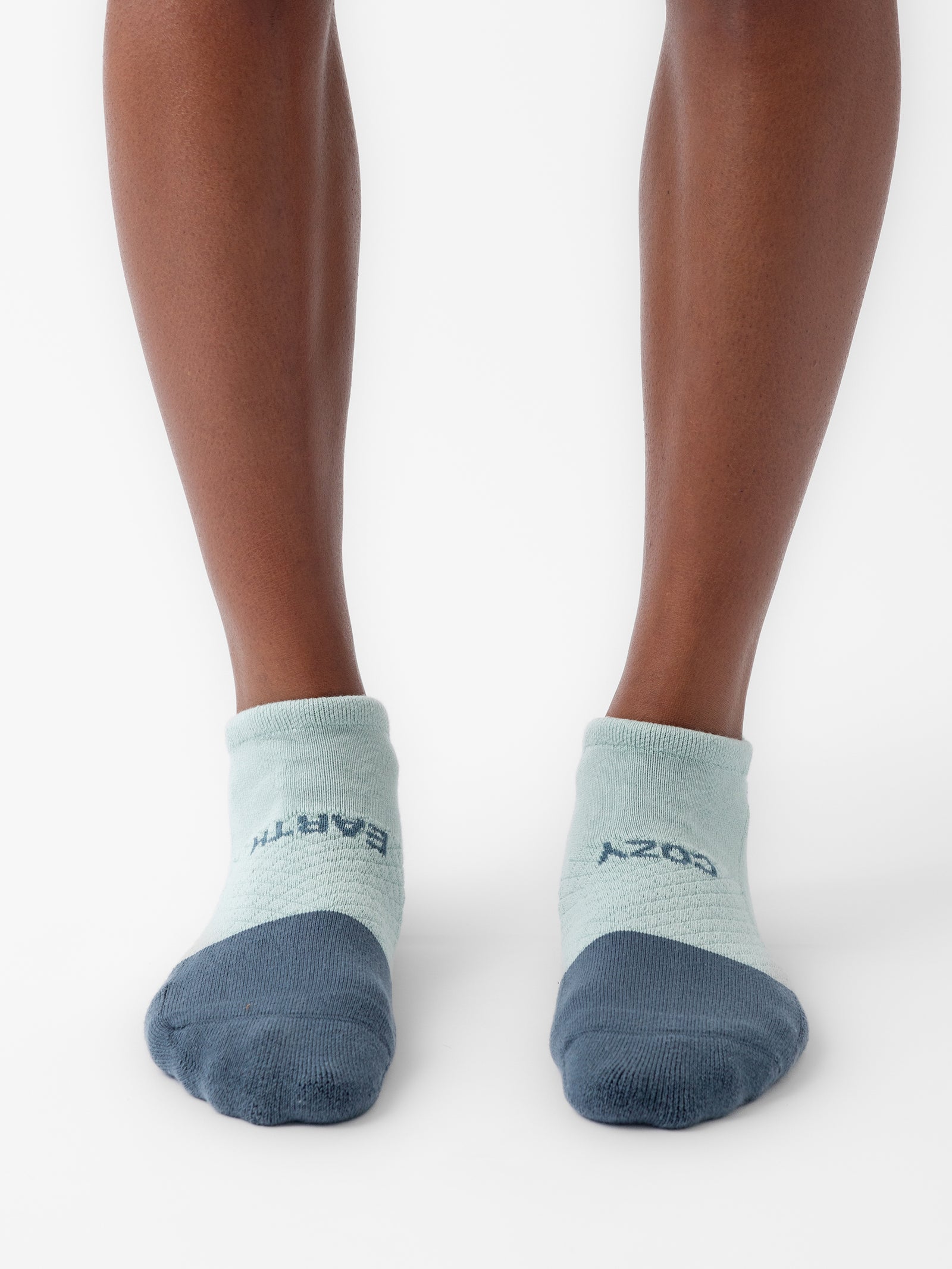 A pair of feet wearing light blue ankle socks from the Cozy Earth Essential Ankle Sock 2-Pack, featuring dark blue toes and heels. One sock displays the word "EARTH" while the other reads "COZY. 