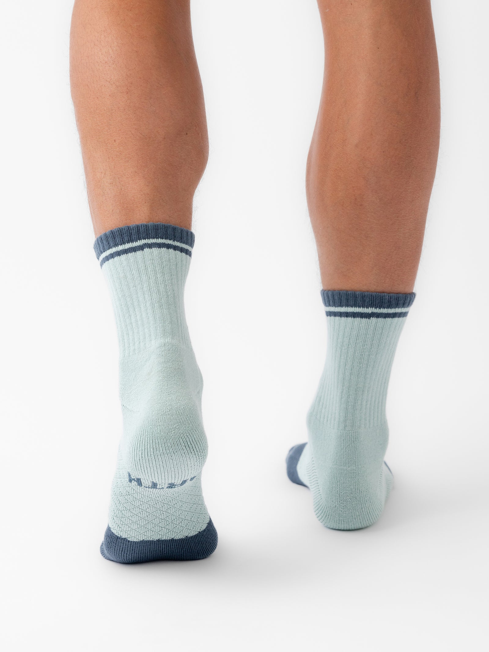 A person's lower legs and feet are shown from the back, wearing Cozy Earth's Essential Quarter Sock 2-Pack in a light blue color with dark blue heel, toe, and cuff accents. The image is set against a white background. 