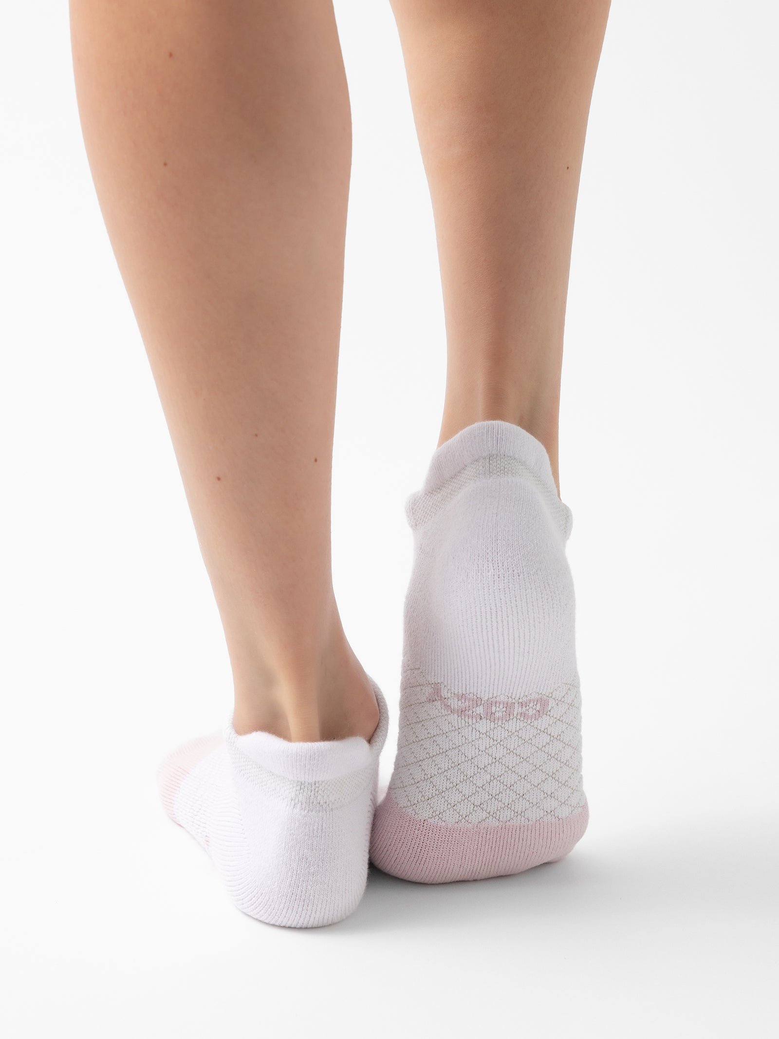 A person standing with their back facing the camera, displaying only their bare lower legs and feet. They are wearing Cozy Earth's Essential Ankle Socks, featuring low-cut white socks with pink heels and soles on a plain white background. 