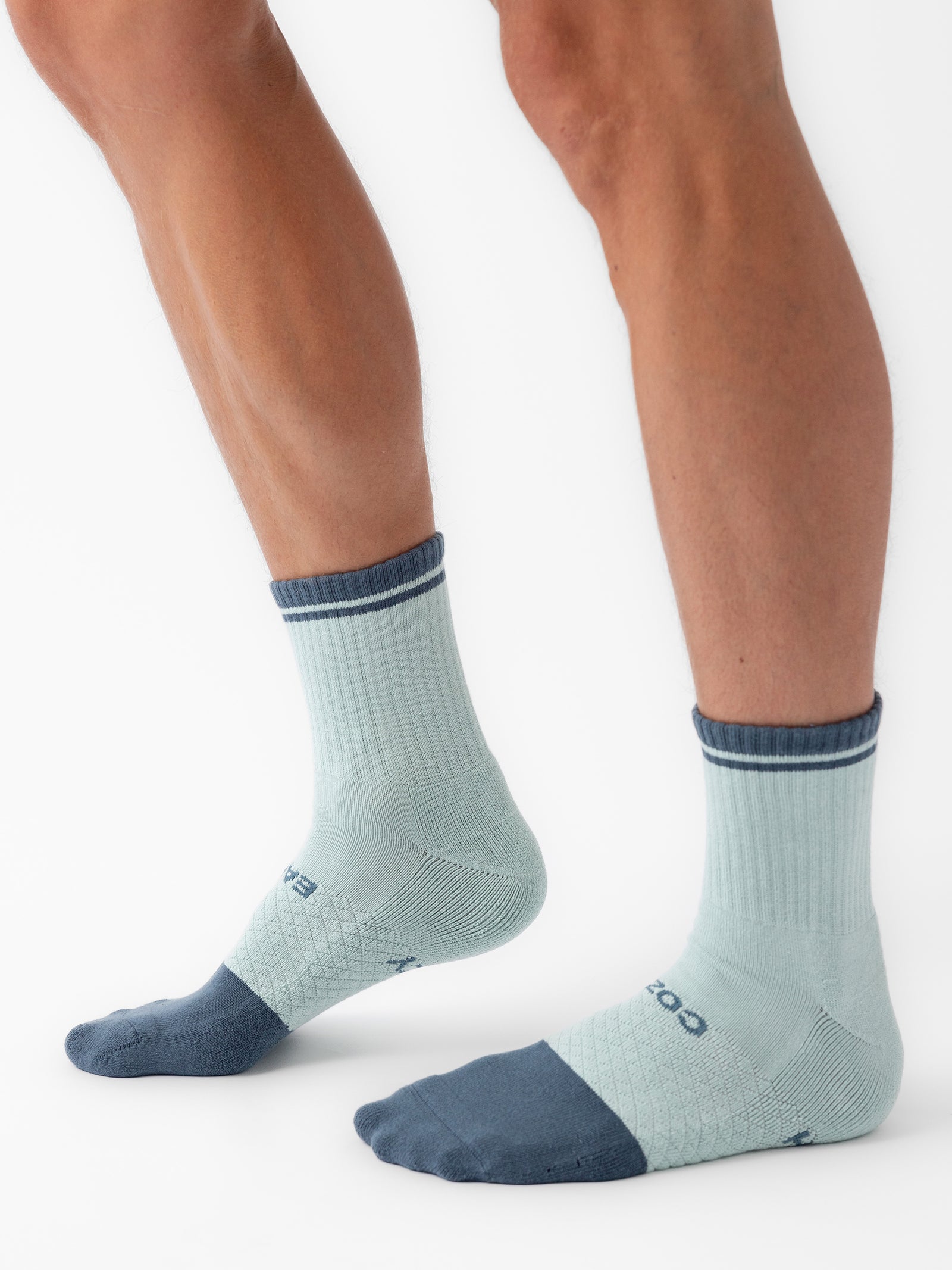 A person is wearing Cozy Earth's Essential Calf Socks in a two-tone color scheme: light blue with darker blue toes, heels, and cuffs. They are standing on a white background, showcasing the socks' crisscross pattern and visible branding near the toes. 