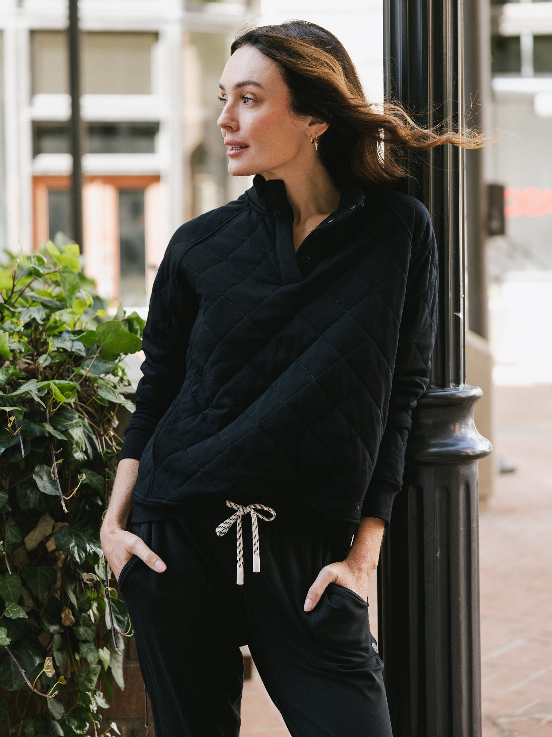 A woman with long brown hair stands outdoors, leaning against a black pole. She is wearing the Cozy Earth Women's Ultra-Soft Bamboo Quilted Snap Pullover and black pants with a white drawstring. Her head is turned to the left as she gazes into the distance, with green foliage visible behind her. 
