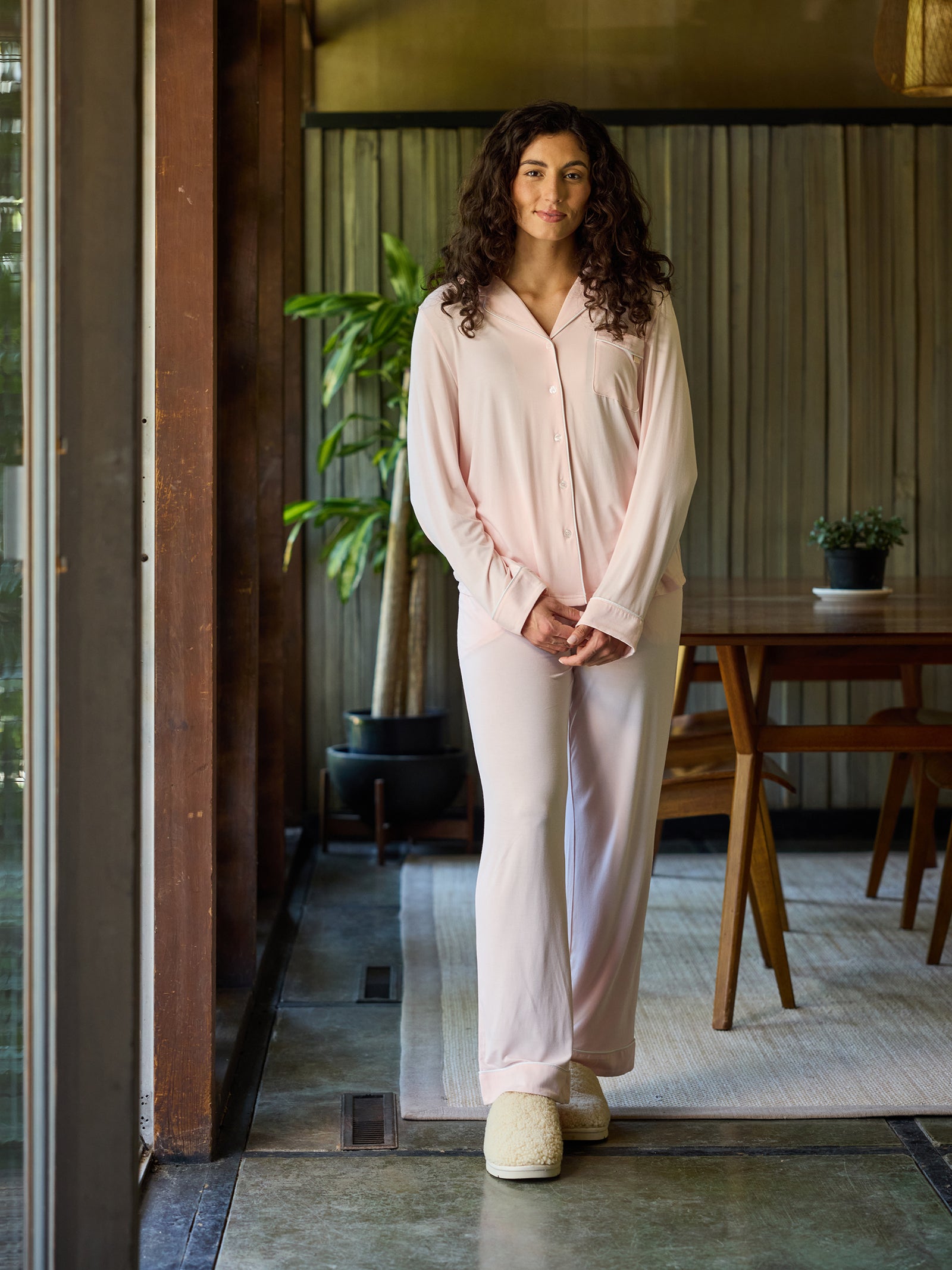 A person with long curly hair is standing indoors near a window, wearing a Women's Long Sleeve Bamboo Pajama Top in Stretch-Knit by HIDE along with light pink pajama bottoms and slippers. Behind them is a wooden dining table with chairs, a potted plant, and a rug on the floor. The room has modern decor with wooden accents and natural light. 