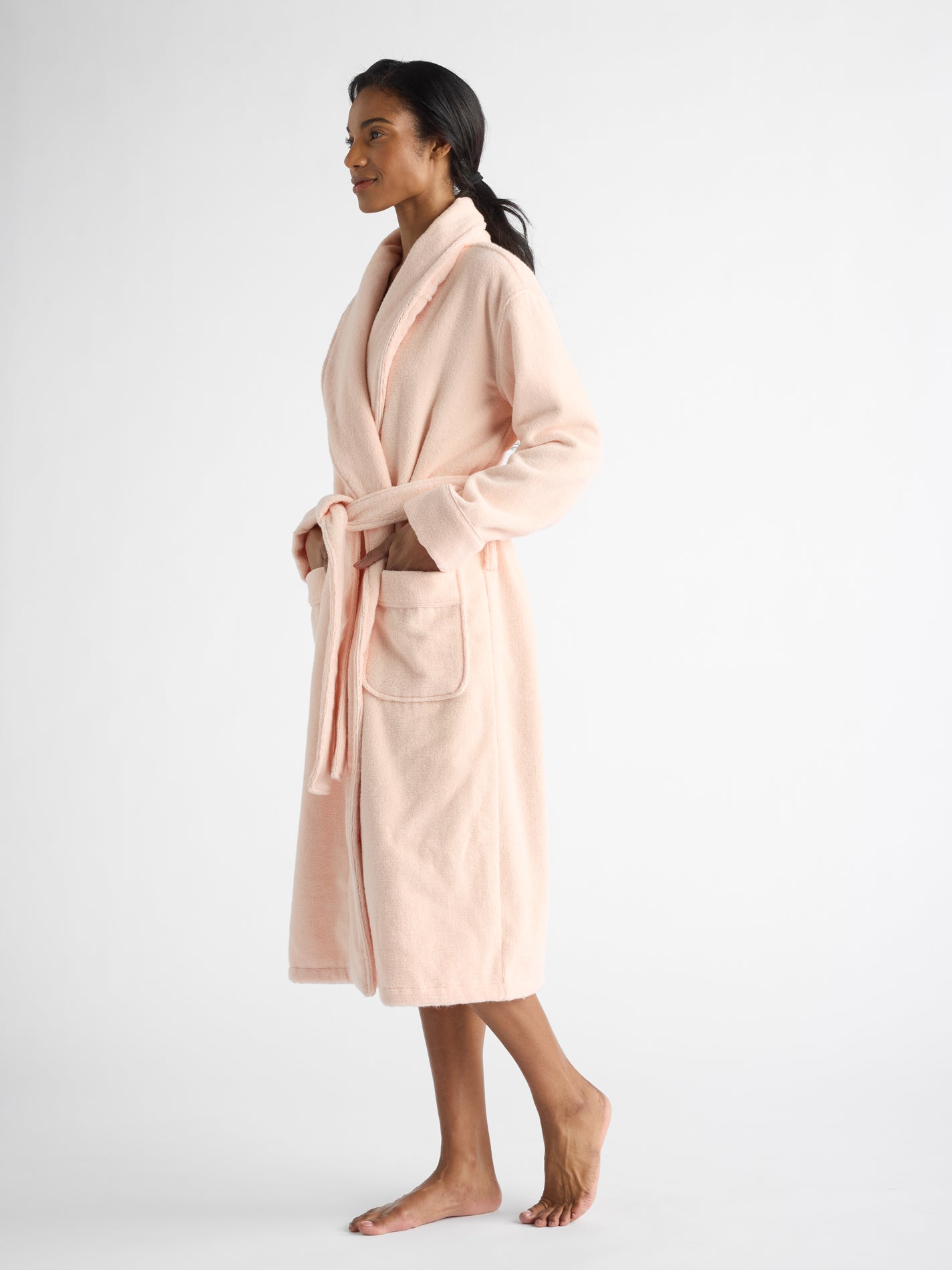 A profile view shows a person in a Cozy Earth's Luxe Bath Robe, pale pink with shawl collar, pockets, and a belt. They stand barefoot on a white background, looking forward with a slight smile. 