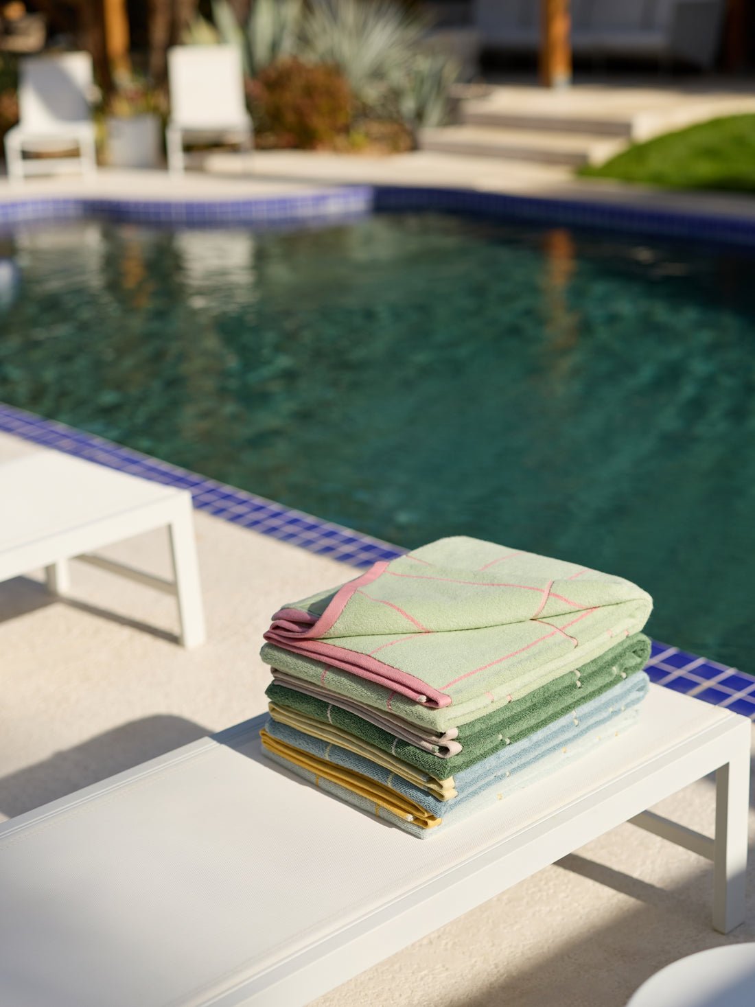 A stack of Cozy Earth's Windowpane Resort Towels in pastel colors sits on a white poolside lounge chair with a blue-tiled pool reflecting sunlight. Another similar chair is partially visible nearby.