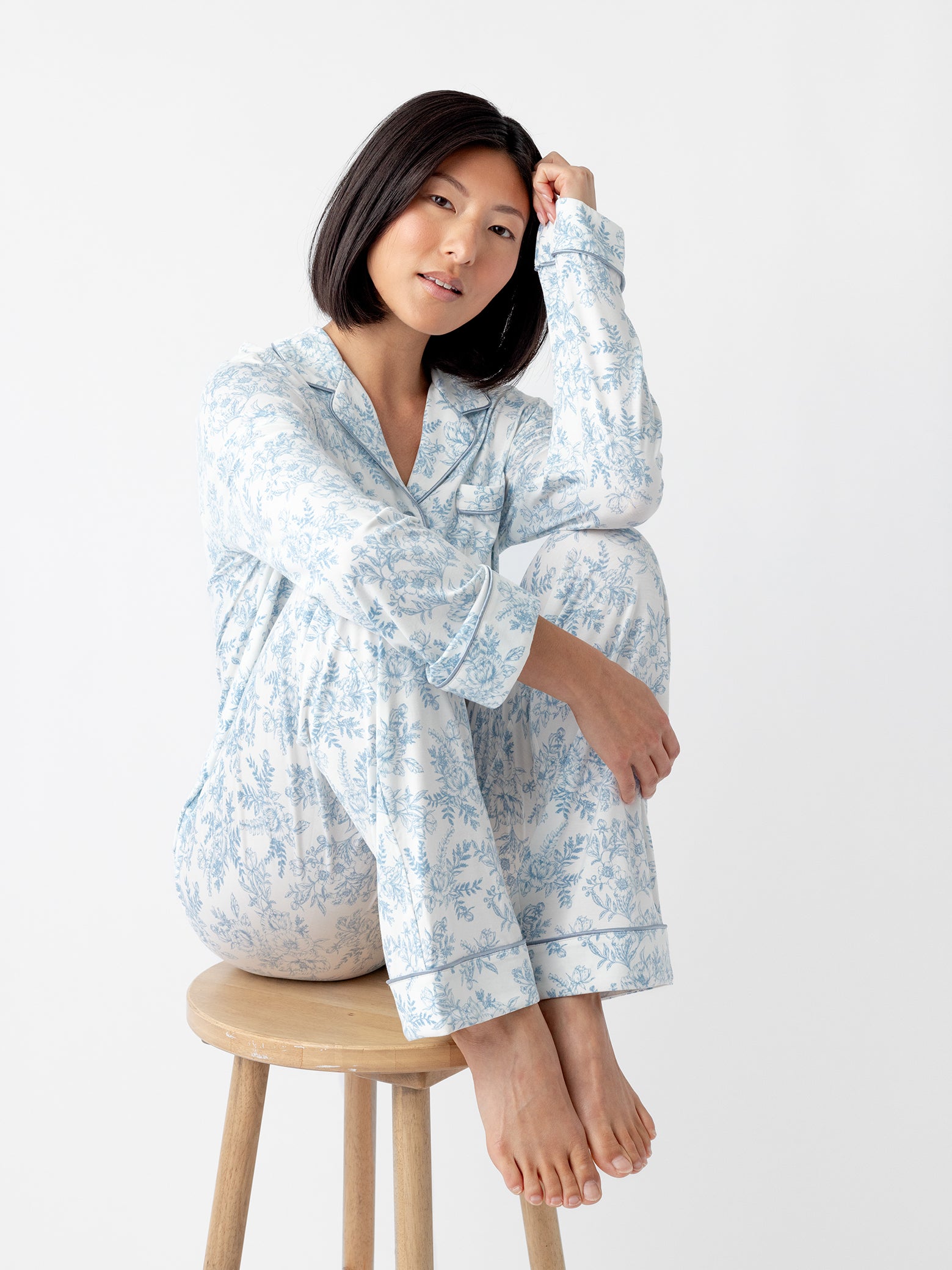 A person with shoulder-length black hair sits on a wooden stool, wearing Cozy Earth Women’s Bamboo Stretch-Knit Long Sleeve Pajama Top in light blue floral pattern. They have a relaxed pose, resting one hand on their knee and the other near their head, against a plain white background. 