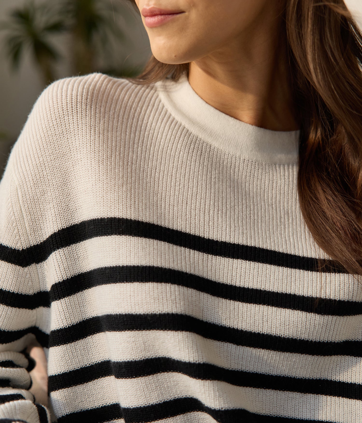 A person wearing the Cozy Earth Women's Rowan Sweater with cream and black stripes stands against blurred foliage, their head is partially visible without a full view of the face. 