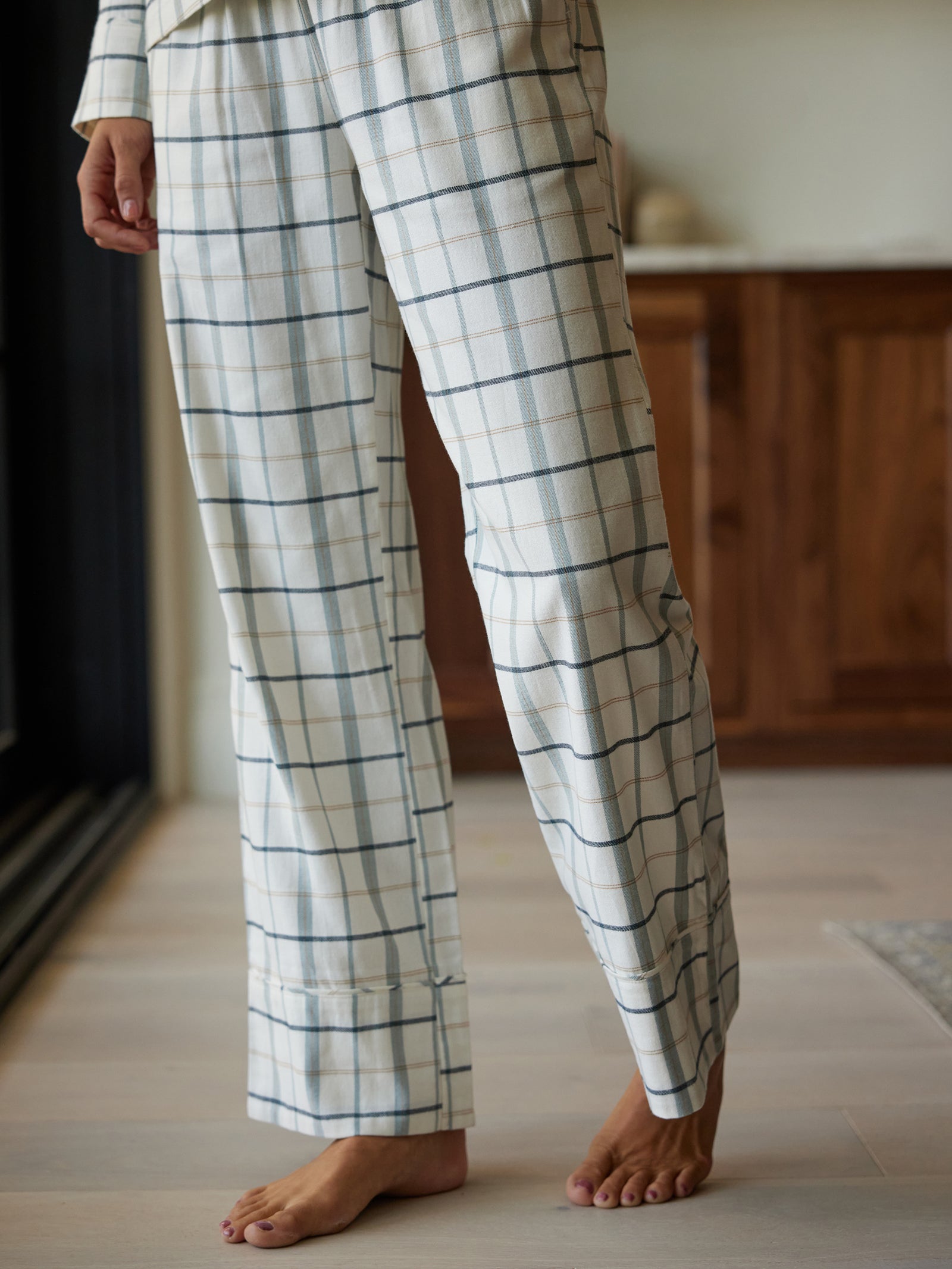 A person stands indoors, wearing Cozy Earth's Women's Farmhouse Pajama Pant, featuring a loose-fitting design with a white plaid pattern accented by thin blue lines. The person's right foot is lightly raised off the ground, and the background includes a wooden cabinet and light-colored flooring. 