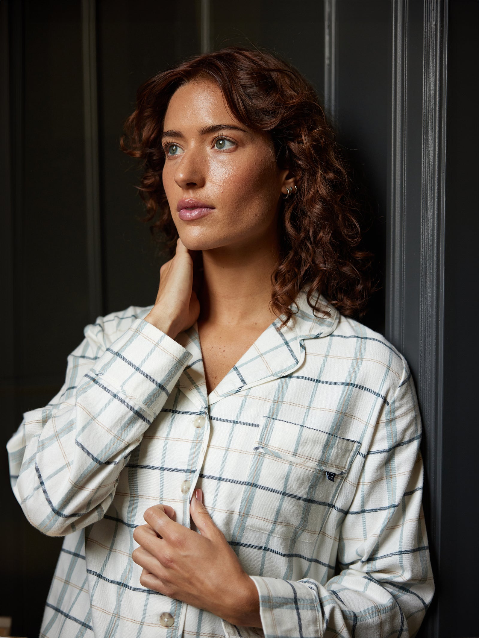 A person with curly brown hair gazes off to the side thoughtfully. They are wearing a Women's Farmhouse Long Sleeve Pajama Top from Cozy Earth in a white and blue checkered pattern, and their left hand is touching their neck. The background consists of dark, blurred vertical panels. 