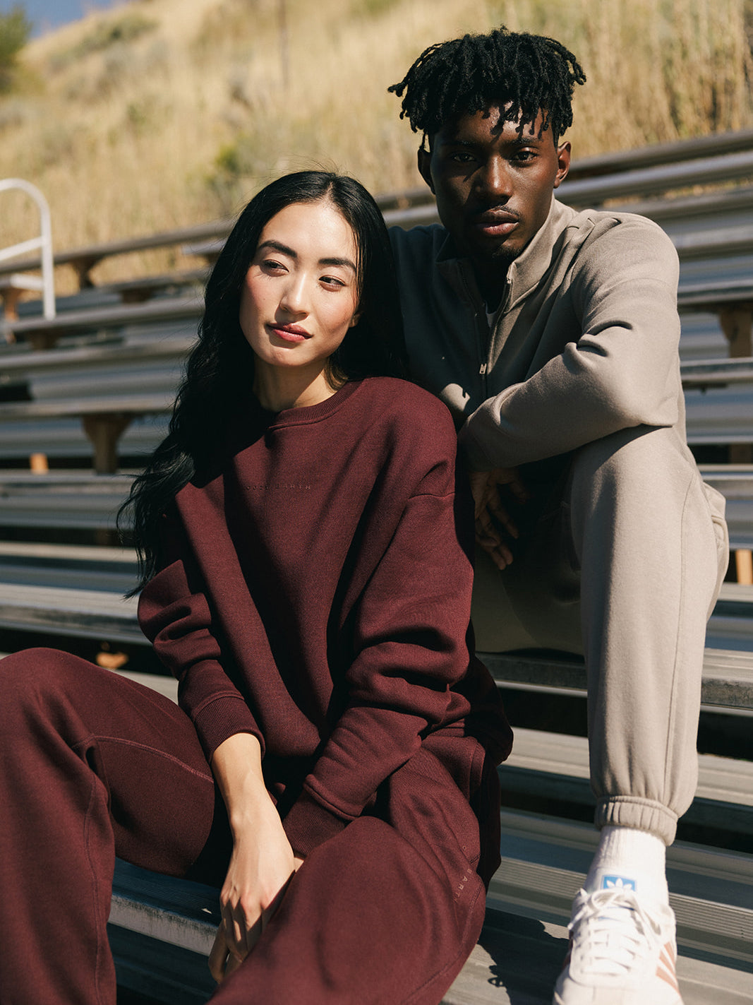 A woman in a Cozy Earth Women's CityScape Crewneck and matching pants sits on bleachers next to a man in a light gray tracksuit. They pose casually in a sunlit outdoor setting with tall grass in the background. 