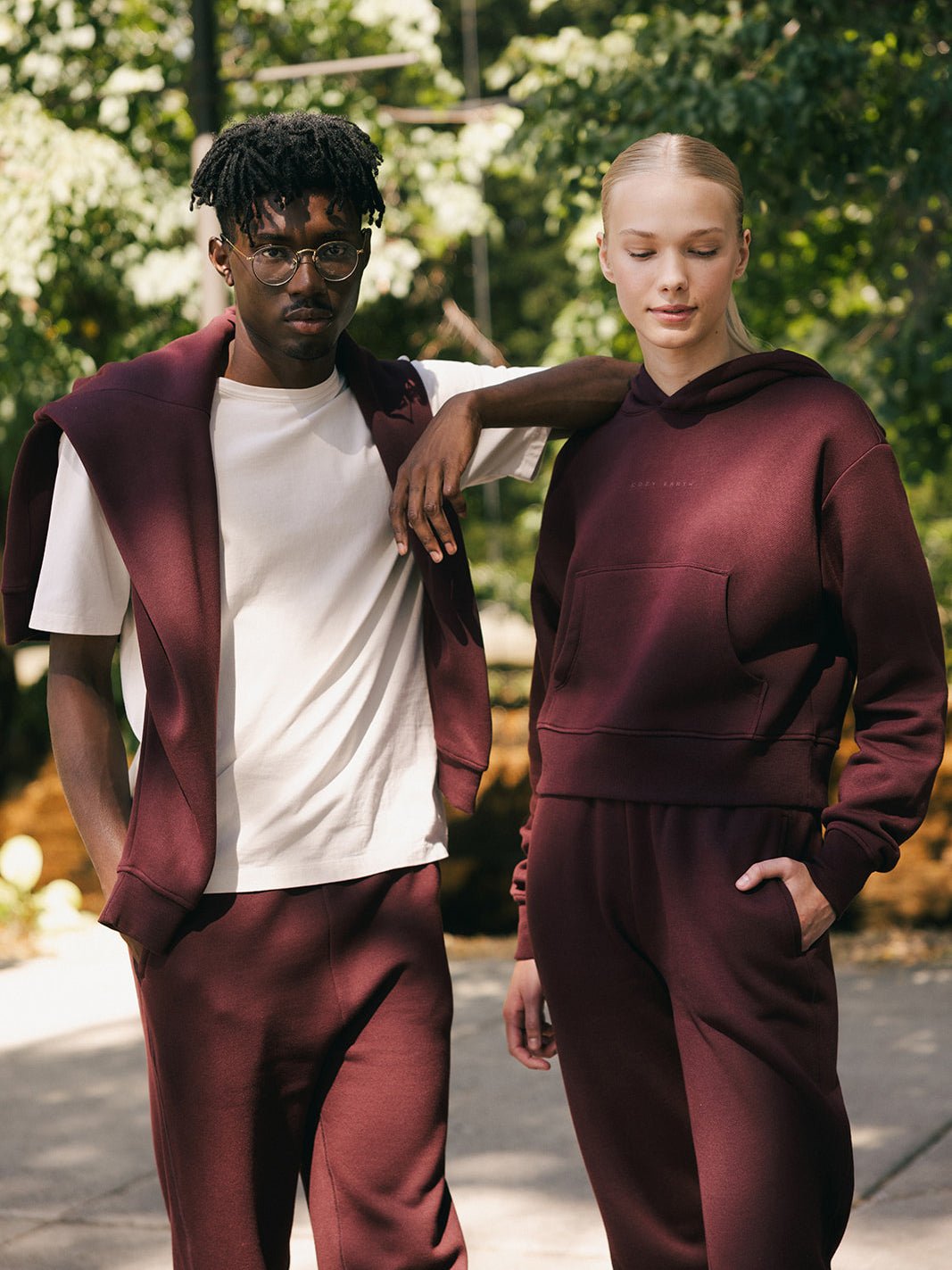 Two individuals stand outdoors in matching burgundy outfits. The man on the left wears glasses, a white t-shirt, and a burgundy sweater draped over his shoulders. The woman on the right sports Cozy Earth's Women's CityScape Cropped Hoodie and matching pants, with one hand in her pocket. They are both gazing at the camera. |Color:Burgundy