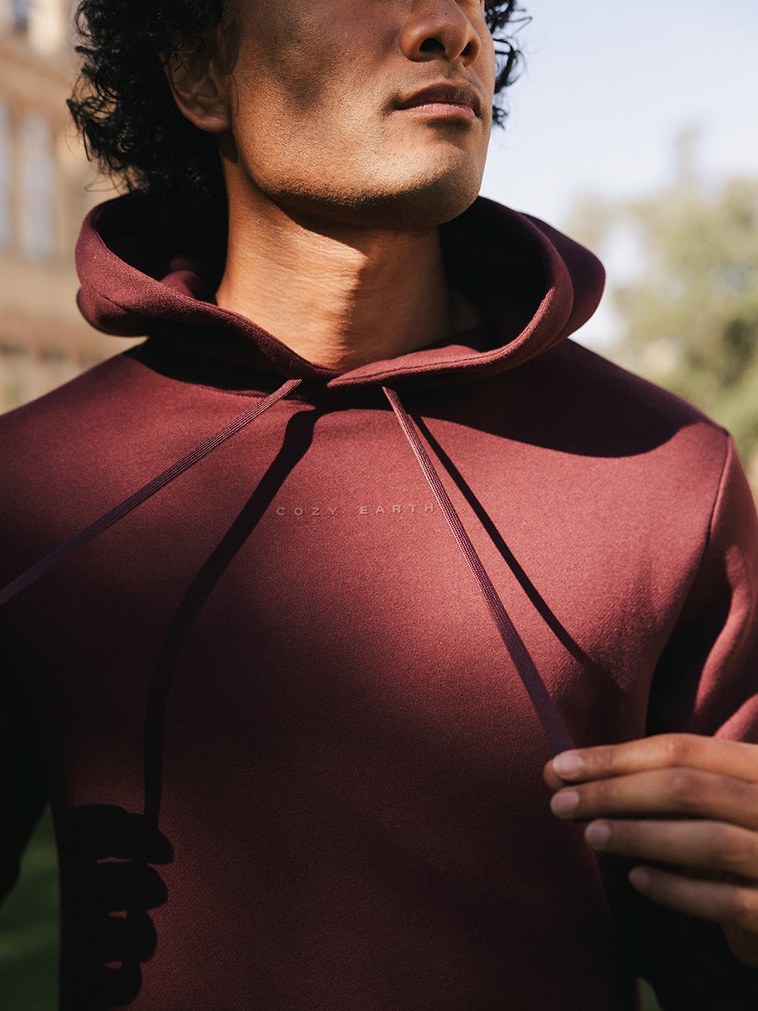 Close-up of a person wearing a Men's CityScape Hoodie in maroon, adjusting the hood strings with both hands. The text "Cozy Earth" is visible on the front of the hoodie. The background shows blurred outdoor scenery with trees and a building. 