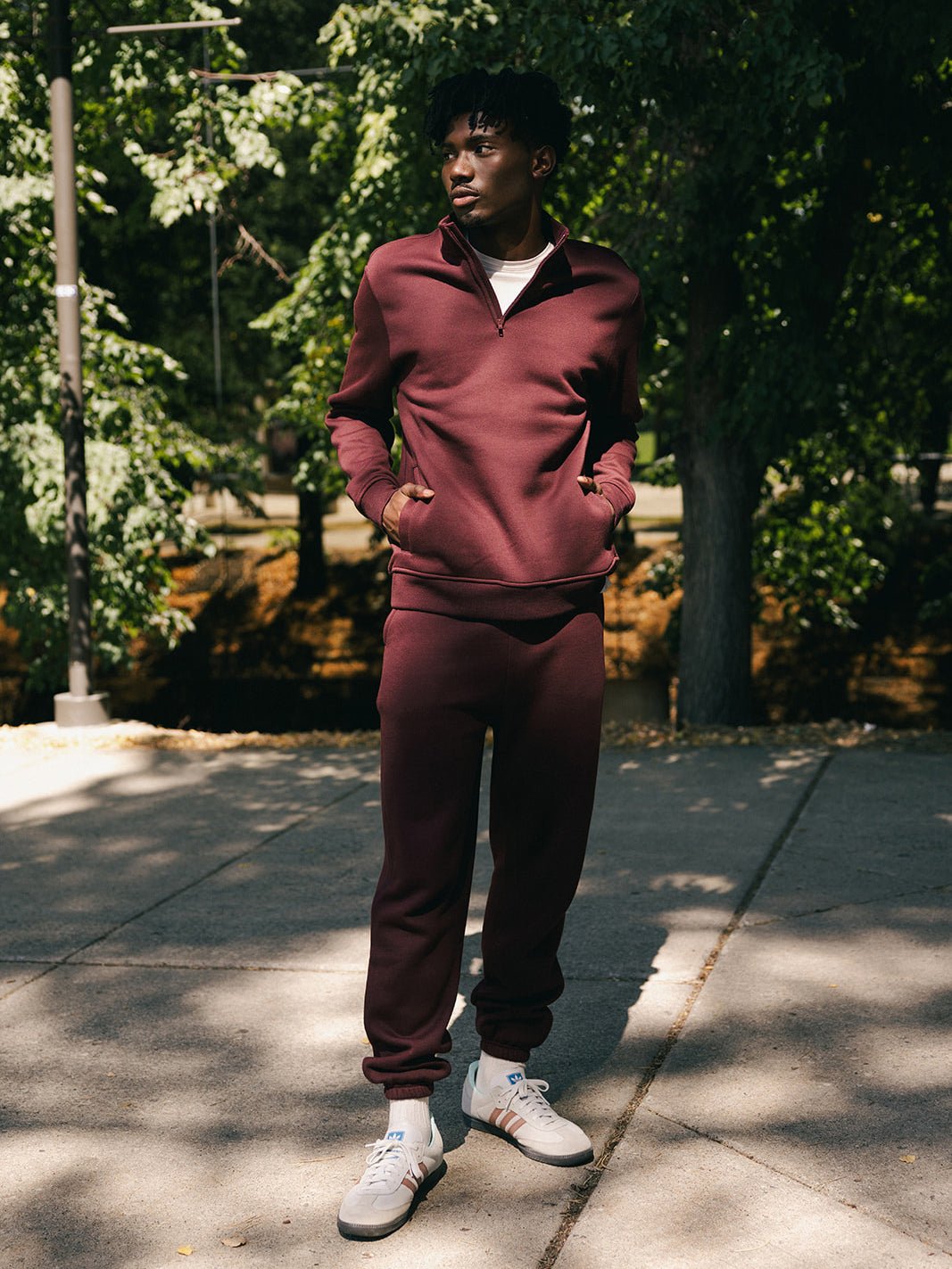 A person stands outdoors on a sunlit sidewalk, wearing Cozy Earth's Men's CityScape Quarter Zip in maroon and white sneakers. They have their hands in their pockets and are looking off to the side. The background features trees with green foliage, casting dappled shadows on the ground. 