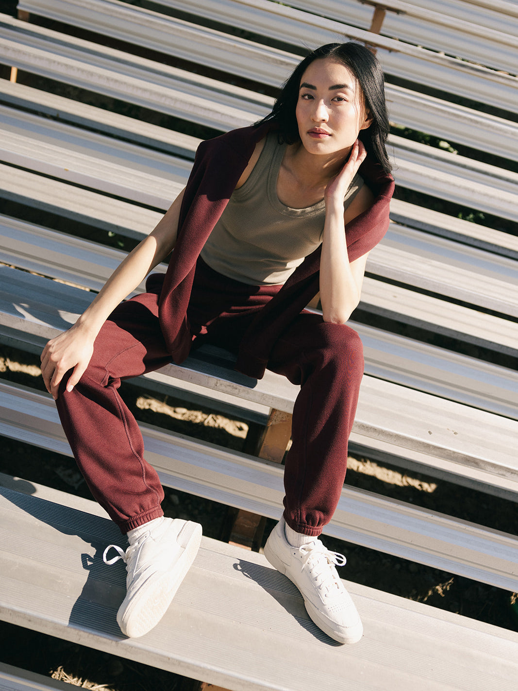 A person with long, dark hair sits on outdoor bleachers, wearing a tan tank top, maroon Cozy Earth Women's CityScape Sweatpant, a matching maroon sweater draped over their shoulders, and white sneakers. The person has a relaxed expression and gazes slightly to their left. 