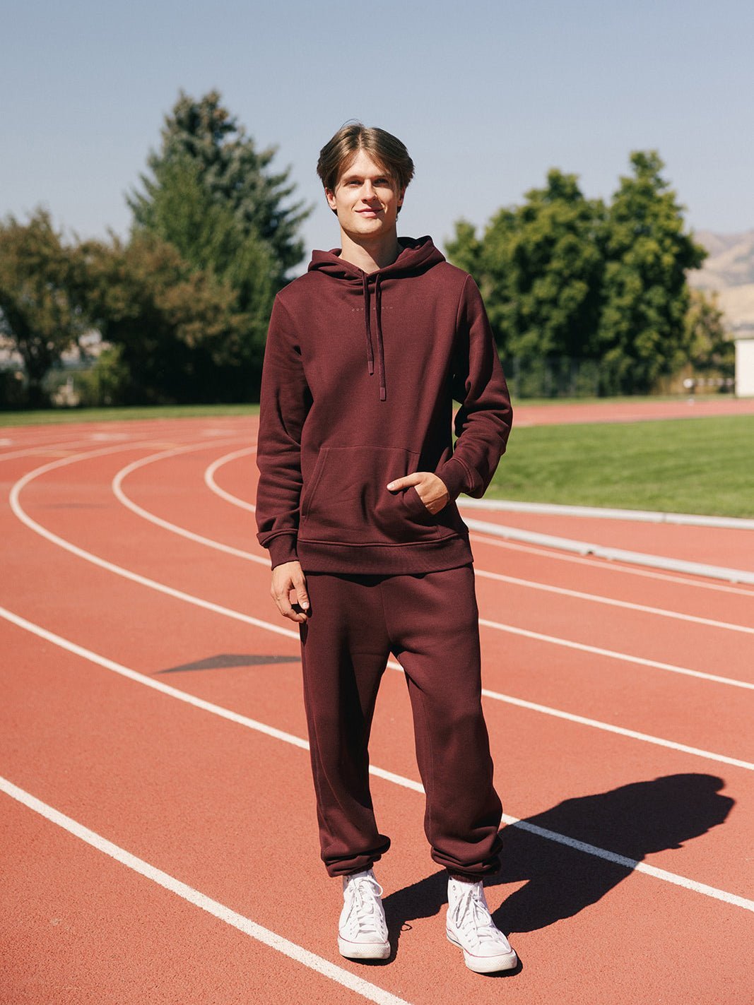 A young man stands on an outdoor running track on a sunny day, dressed in Cozy Earth's Men's CityScape Sweatpants and matching burgundy hoodie, paired with white sneakers. Green trees and a clear sky can be seen in the background. He is smiling and facing the camera. 