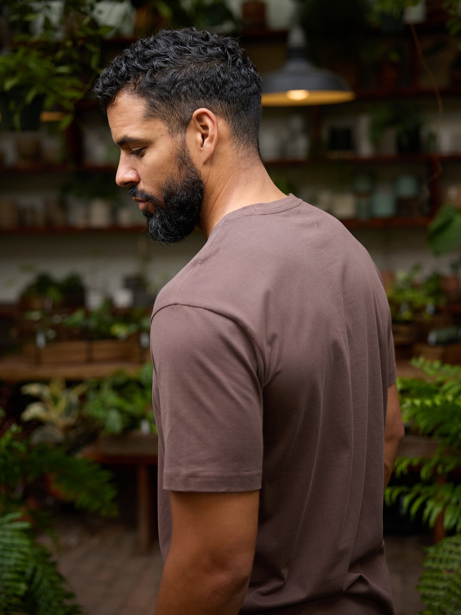 A man with short curly hair and a beard, wearing the Cozy Earth Men's All Day Tee in brown, stands in profile in an indoor garden setting. He is looking downward, surrounded by green plants and rustic shelves with various items in the background. The atmosphere is serene and natural. 