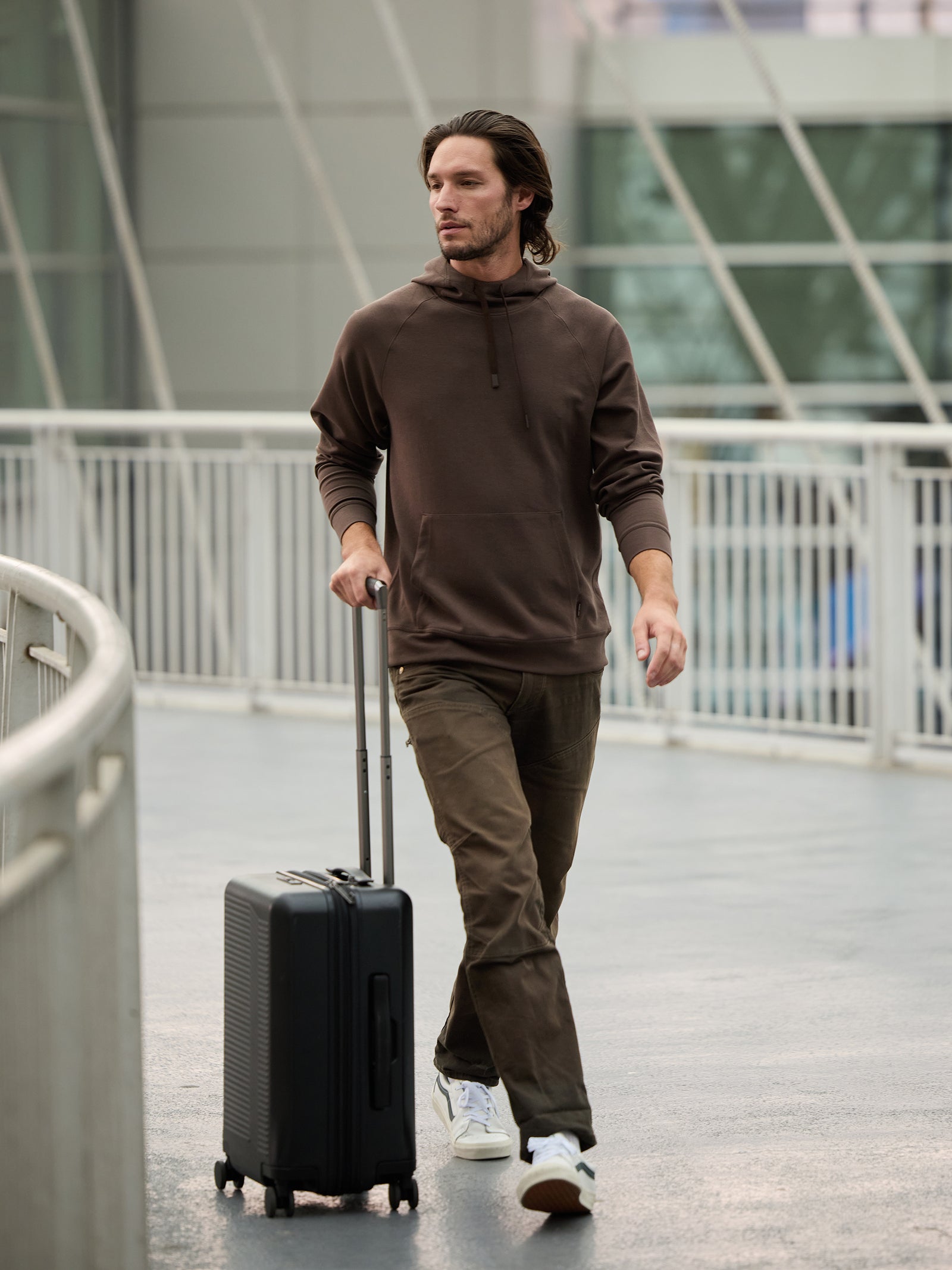 A man in a Cozy Earth Men's StretchTech Hoodie and olive green pants walks on a walkway pulling a black rolling suitcase. He has shoulder-length hair and is wearing white sneakers. The background features a modern building with a glass exterior. 