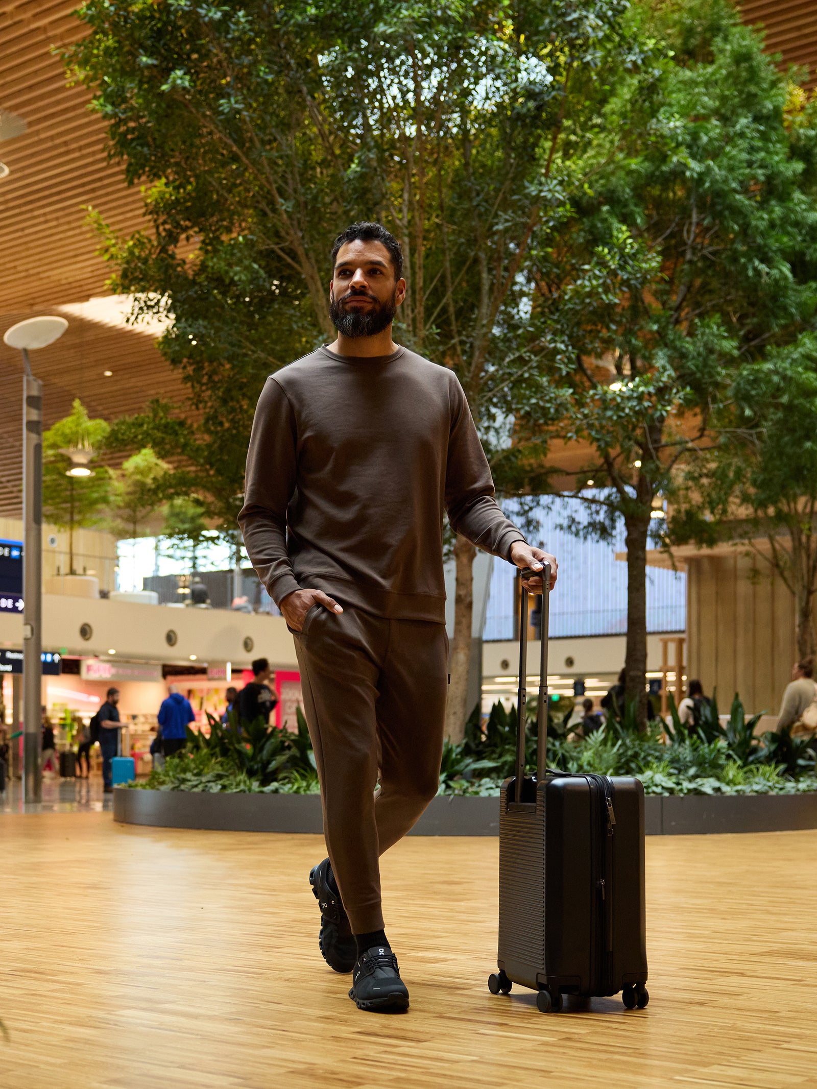 A man with a beard, dressed in a Cozy Earth Men's StretchTech Jogger and black sneakers, walks through an indoor space pulling a dark rolling suitcase. The background features lush green trees, a wooden floor, a high ceiling, and a few people and stores visible in the distance. 