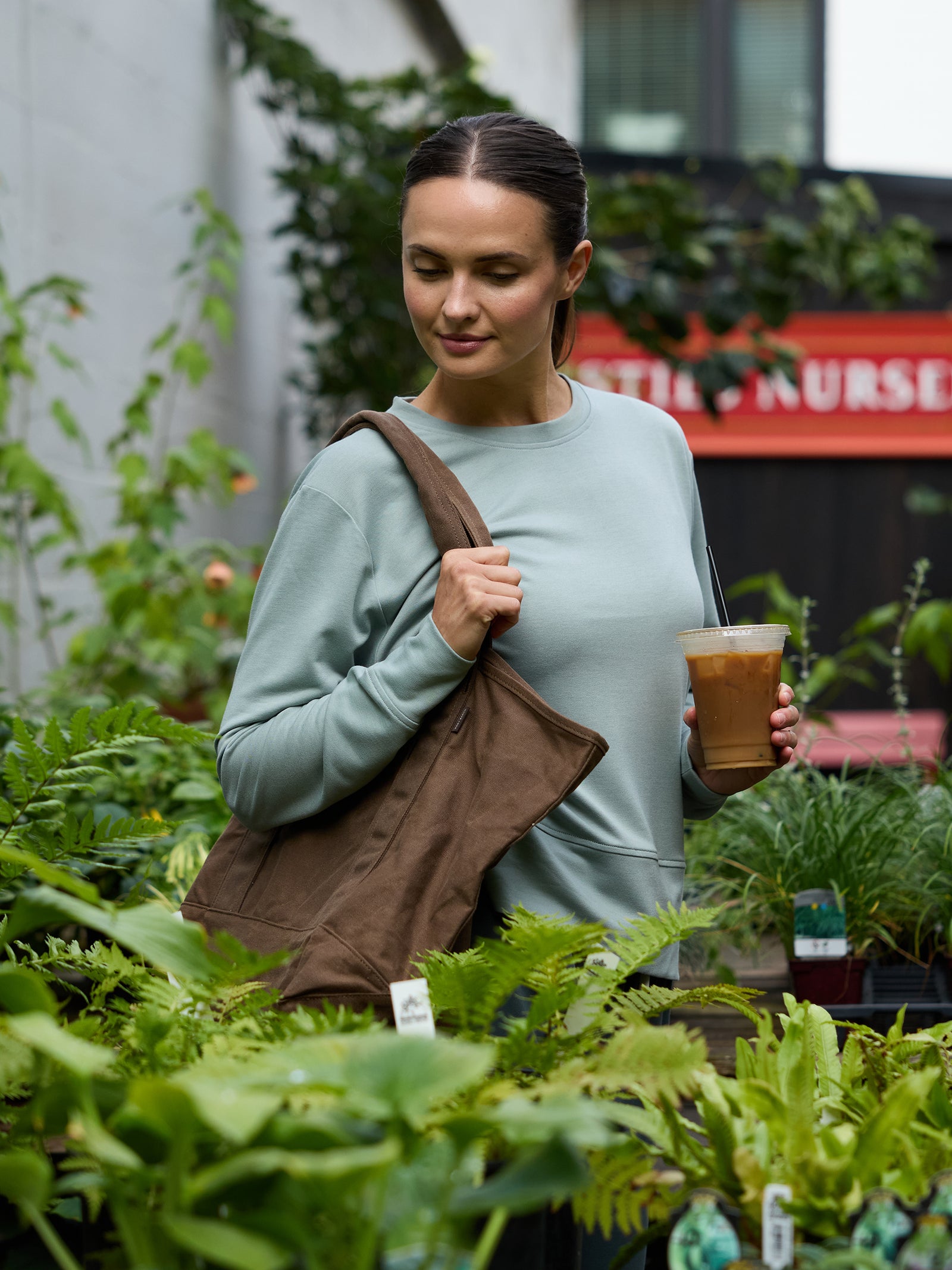 A person with long hair tied back, wearing a light green sweater and carrying a Cozy Earth Waxed Canvas Tote, is holding an iced coffee. They are standing in a lush plant nursery, surrounded by various green plants and ferns. A blurred sign in the background reads "NURSERY. 