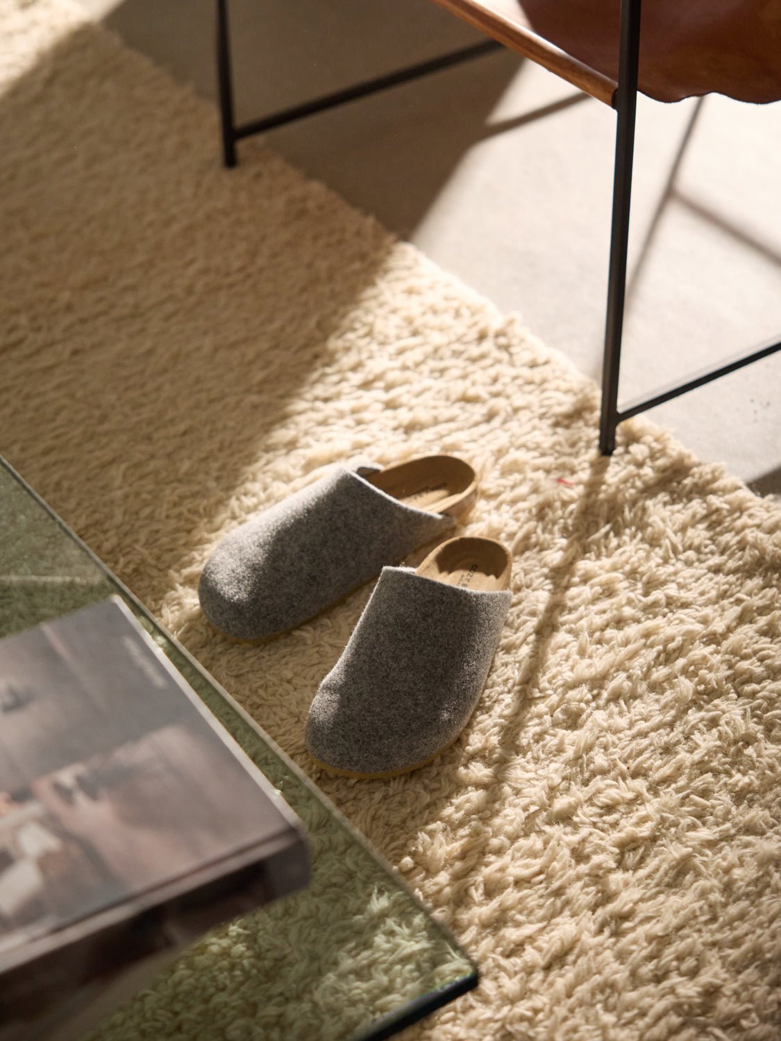 A pair of Cozy Earth's Lakehouse Clog wool slippers rests on a soft, cream-colored shag rug near a glass coffee table and leather chair. Sunlight from a nearby window casts warm shadows across the scene. 