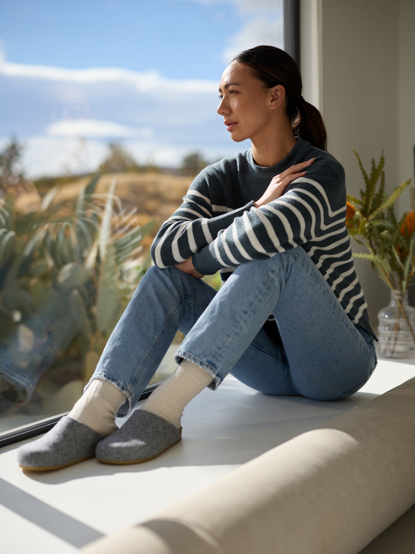 A person sits on a window seat, gazing outside thoughtfully. They are wearing a striped sweater, jeans, and Cozy Earth's Lakehouse Clog slippers. Sunlight illuminates the scene, with plants and sky in the background. 