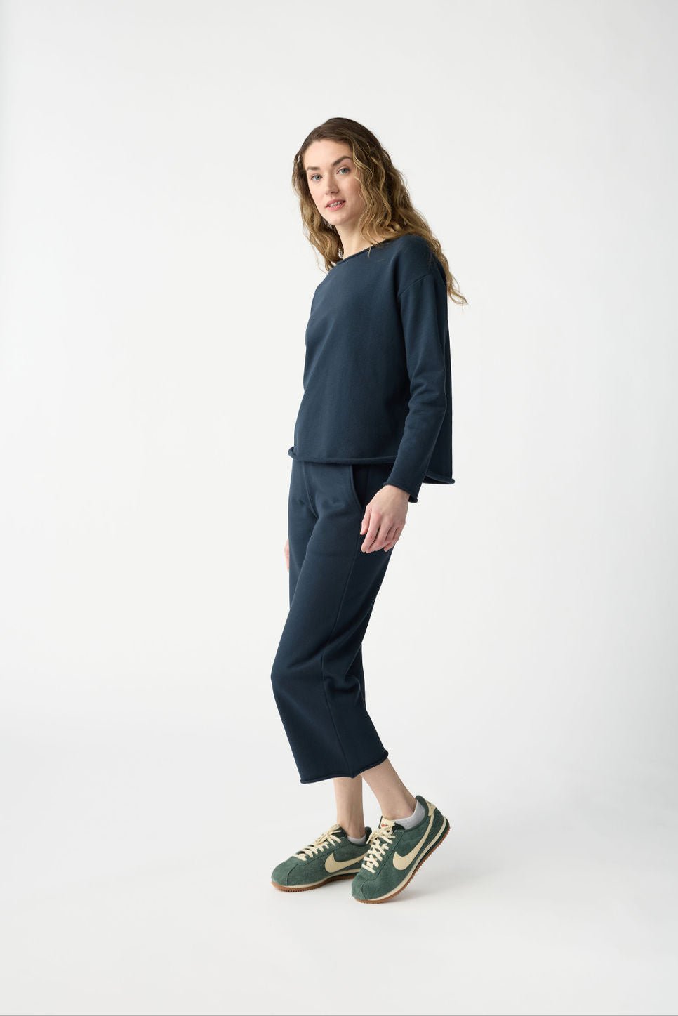 A person with long, wavy hair is wearing the Women's Chelsea Set from Cozy Earth with green sneakers. They stand on a plain white background, looking slightly to the side in a relaxed posture. 