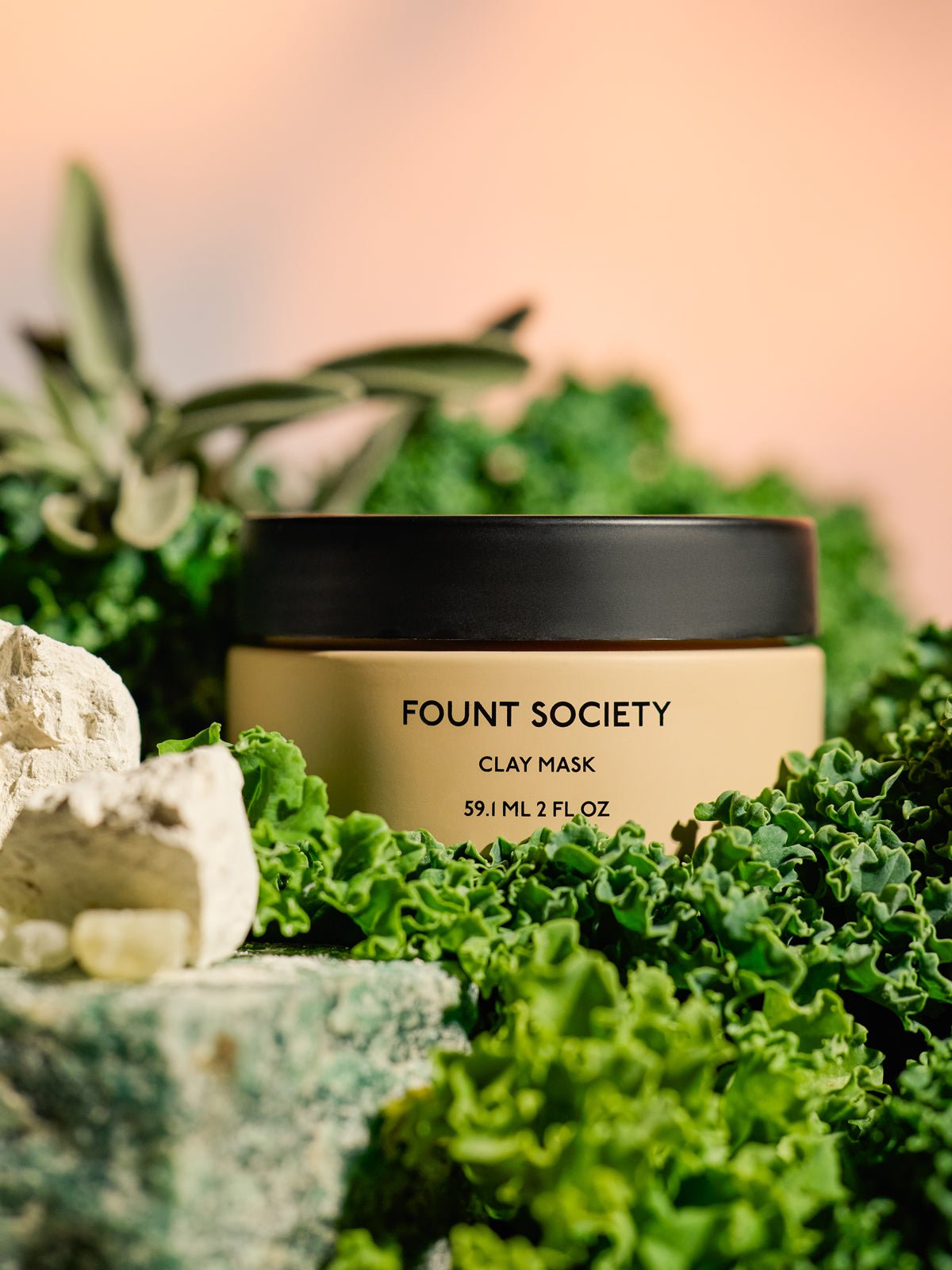 A Cozy Earth Clay Mask container is nestled among lush greenery and rocks, with a blurred pastel background. 