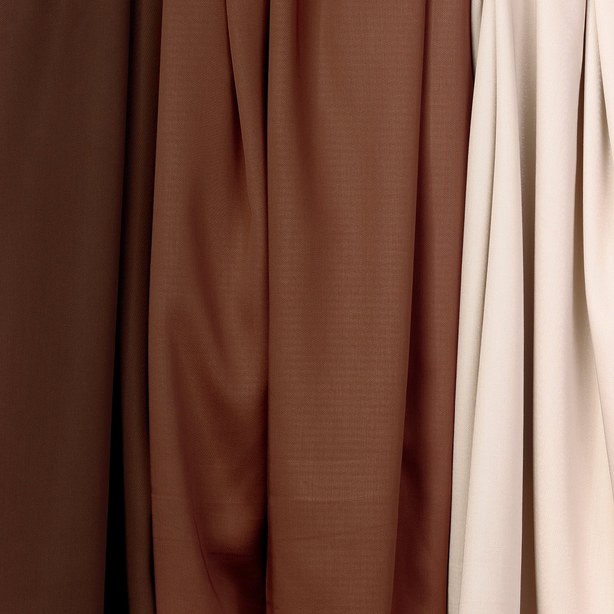 Close-up of various drapes from the Bamboo Sheet Set by Cozy Earth hanging side by side. The fabrics display a gradient of brown hues, transitioning from dark brown on the left, to medium brown in the center, and an off-white or beige shade on the right. The texture is smooth and lightweight. 